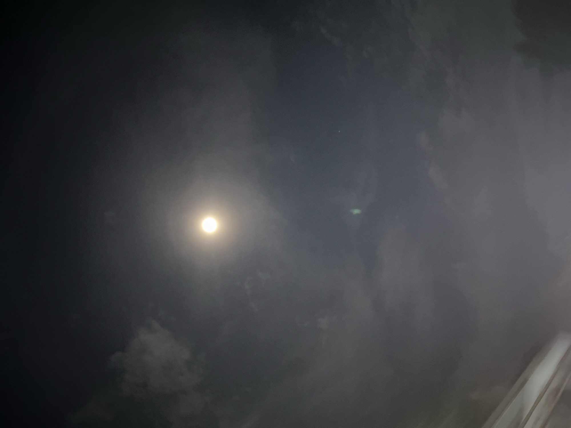 The image depicts a night sky illuminated by a bright, full moon partially obscured by clouds. The clouds are wispy and diffuse, creating a soft glow around the moon. The overall atmosphere is dark and moody, with hints of luminescence in the celestial bodies. There are no discernible human figures or man-made structures in the frame, suggesting an outdoor setting. The bottom portion of the image shows a sliver of what appears to be an artificial structure, possibly a building or a bridge, with diagonal lines indicating its form. This detail, however, is largely out of focus and does not detract from the prominence of the night sky and moon. The scene suggests a quiet, perhaps even serene, moment under the night sky, with the natural beauty of the moon and clouds taking center stage. Without further context or information about the location in Ceurih, Indonesia, it is difficult to provide specific details about the environment beyond what is visible in the image itself. The photograph appears to be taken from a ground-level perspective, looking upwards towards the heavens.