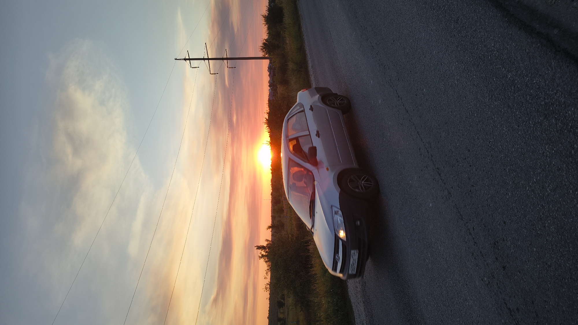 A silver sedan is driving down a rural asphalt road during sunset. The sky is a gradient of orange, pink, and blue, with wispy clouds. Power lines run horizontally across the sky, supported by a tall utility pole on the right side of the image. The car has its headlights on, and the interior lights are also on, illuminating the driver and a passenger. The landscape on either side of the road is covered in tall grass and scattered trees. The car is in motion, as indicated by the slight blur on its wheels and the light trails from its headlights. The overall scene depicts a journey through a picturesque rural setting at dusk.