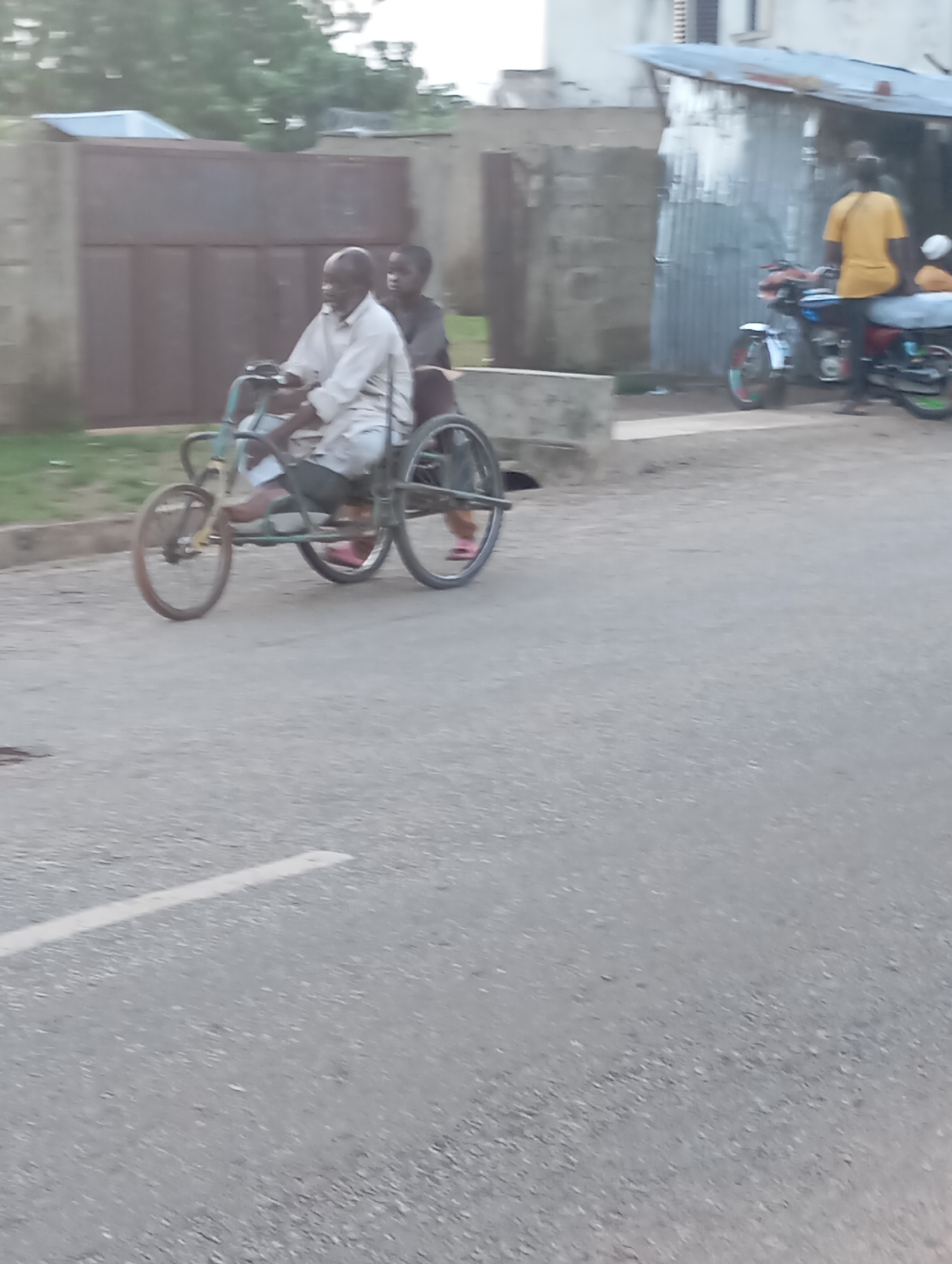 The image depicts a tricycle with two individuals on a paved road in Shendam, Nigeria. An elderly man with a light-colored shirt is operating the tricycle, and a younger person, possibly a child, is seated behind him. The tricycle has a single front wheel and two rear wheels. In the background, there is a building with a corrugated metal roof and a large, dark-colored gate. A few motorcycles are parked near the building. The overall environment suggests an outdoor, possibly semi-urban setting.