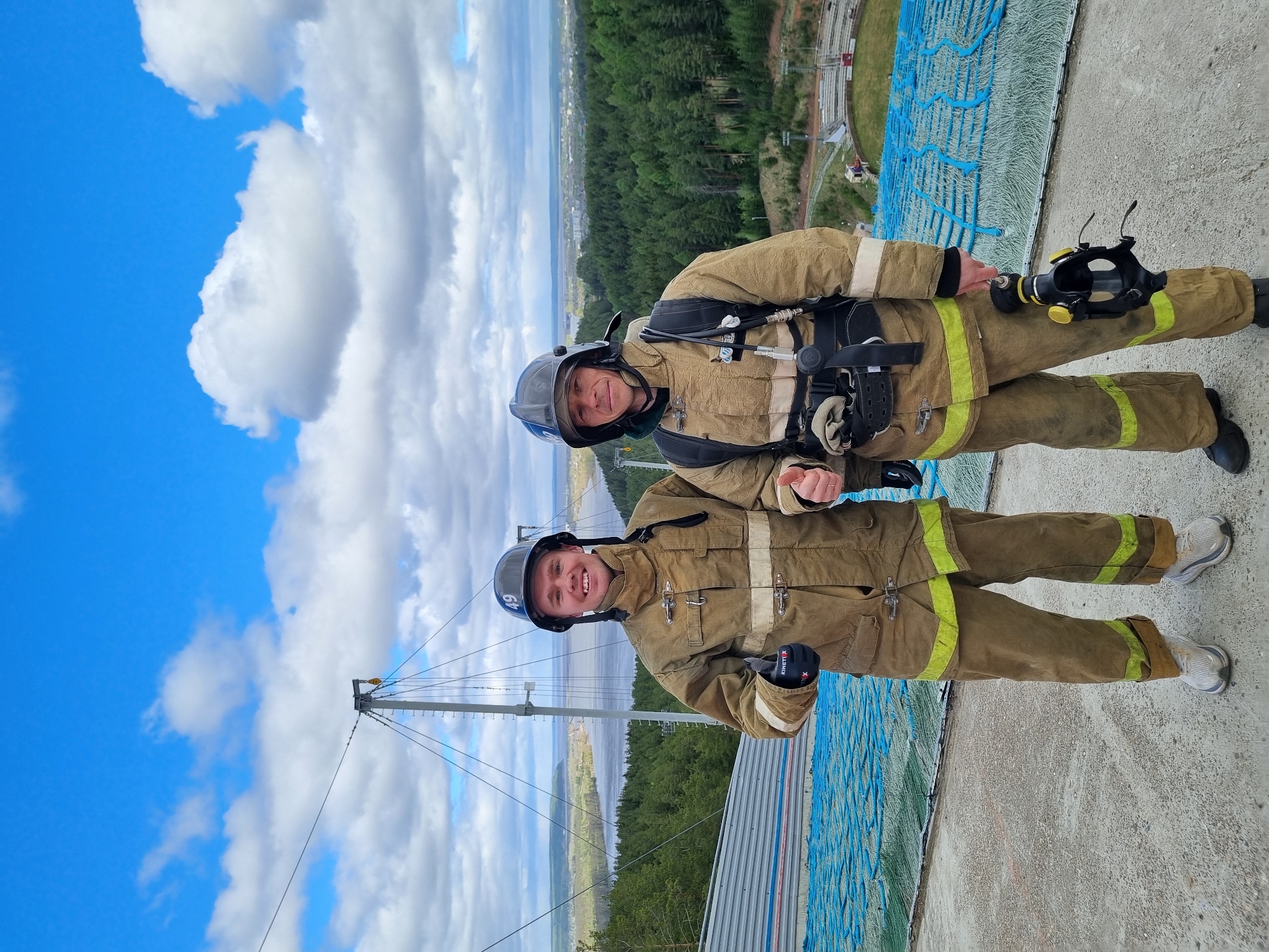 Two individuals dressed as firefighters in tan uniforms and helmets, with breathing apparatus visible on the upper figure, stand on a concrete ledge with a ski jump structure to their right. The background features a cityscape partially obscured by trees and water, with a bright blue sky and white clouds dominating the upper left corner of the image. The location is Прикамский, Russia.