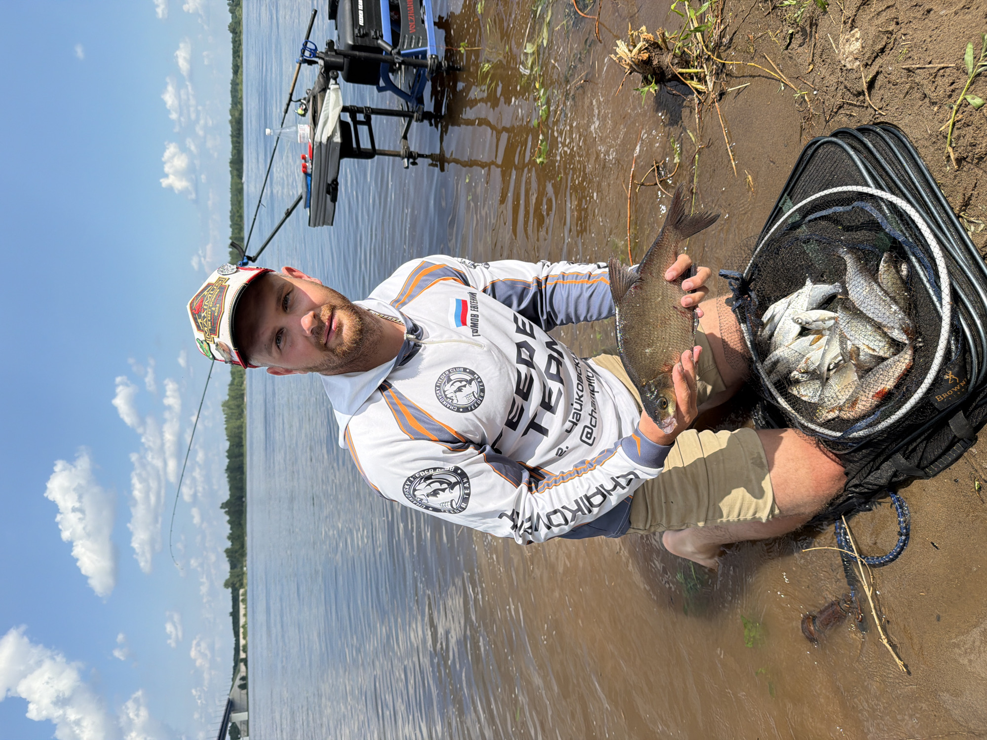 A Caucasian male wearing a white and orange long-sleeved fishing shirt, tan shorts, and a red and white baseball cap is kneeling by the water's edge on a sandy bank. He is holding a single, medium-sized fish in his right hand. In front of him is a black fishing net containing several smaller fish. The man has short brown hair and a beard. The background features a wide river under a bright blue sky with scattered white clouds. The riverbank on the opposite side is lined with trees. Fishing rods are visible in the background, along with a fishing station. The image appears to have been taken in Tarasovo, Russia, likely on a sunny day, with the water reflecting the sky and surrounding environment.