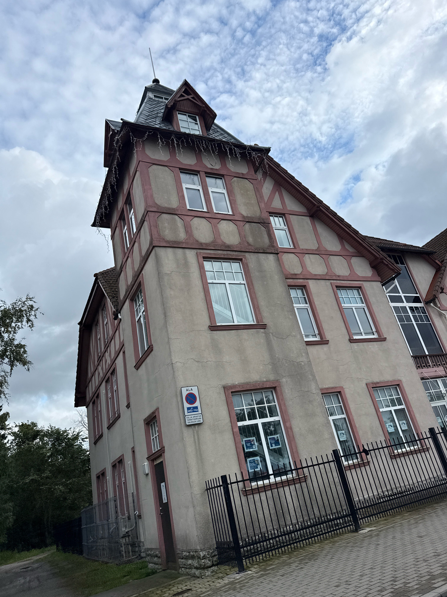 A pale beige, multi-story building with dark red trim and a dark grey, mansard roof stands at an angle under a cloudy sky. The building has multiple white-framed windows of various sizes, some adorned with posters. A dark metal fence runs along the front of the building, bordering a grey cobblestone pathway. To the left of the building, there is a dense green tree and a gravel path. A sign with a blue circle and a red diagonal line over a red boat icon is mounted on the building's exterior. The building appears to be in an urban or suburban setting in Tallinn, Estonia.