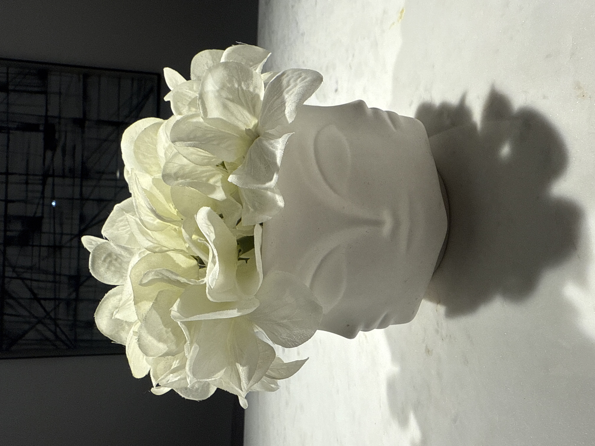 A white vase in the shape of a human face, filled with white artificial flowers, is placed on a white surface. A shadow is cast to the right of the vase. A black framed window with a criss-cross pattern is visible in the background to the left. The image was taken in Loures, Portugal.