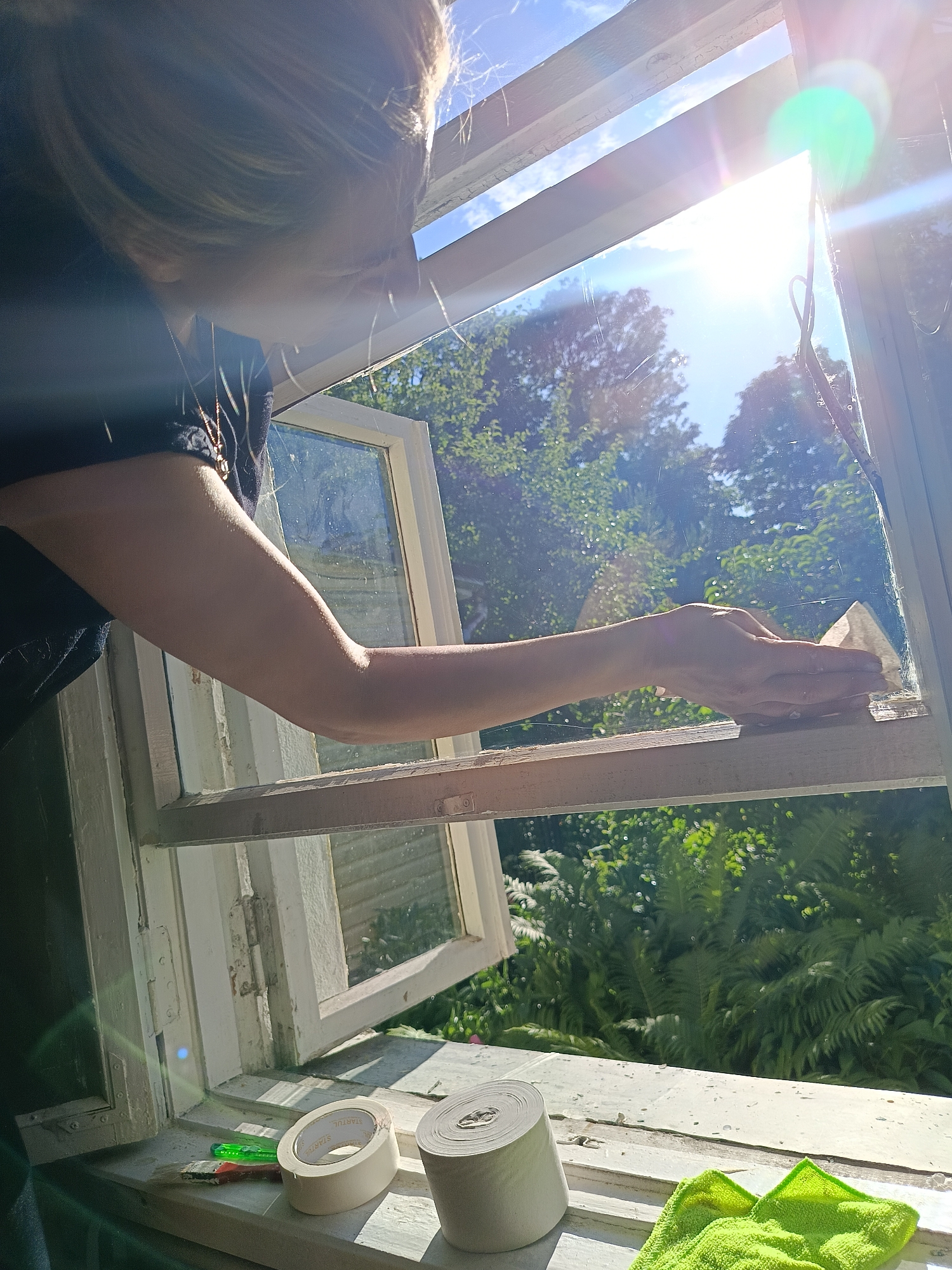 A person's arm and shoulder, wearing a dark shirt and a necklace, are visible from the side as they lean over an open window and clean the frame with a light-colored object. Outside the window, lush green trees and foliage are visible under a bright blue sky with the sun shining, creating lens flares. On the windowsill inside, there is a small paintbrush with red and green handles, a roll of white tape, and another roll of what appears to be paper towels or more tape. A bright green microfiber cloth is also on the windowsill. The window frame is old and painted white, with some peeling paint and visible hinges. The scene appears to be outdoors, possibly in a rural setting or a backyard in Niasvizh, Belarus, given the greenery and the style of the window.