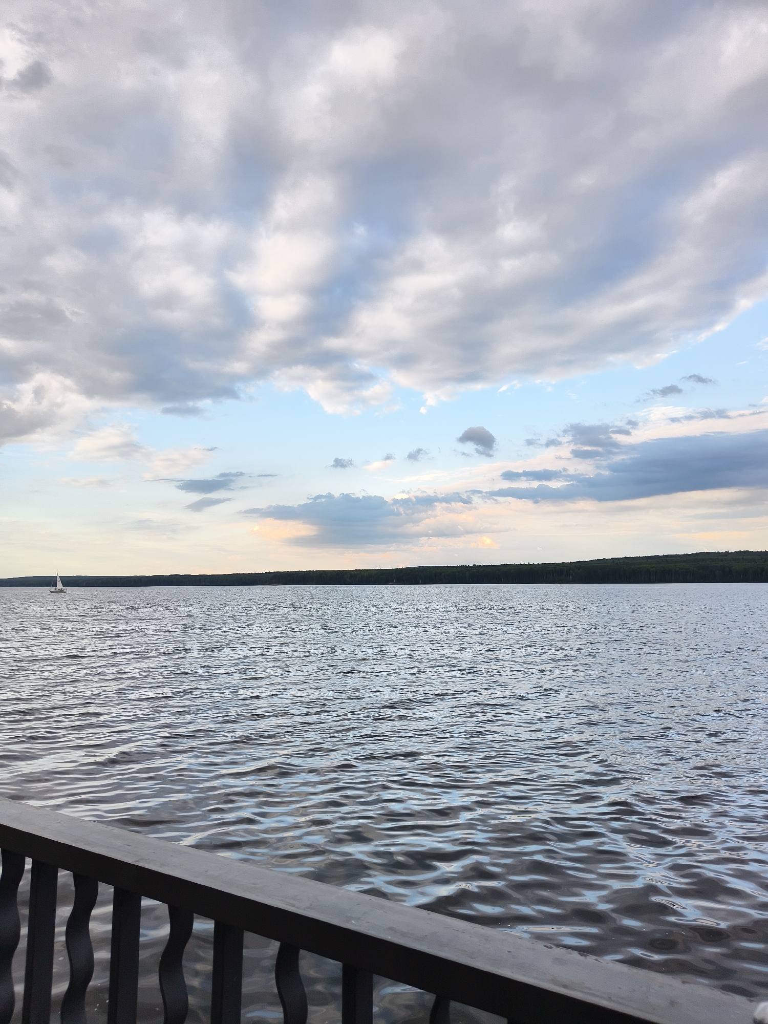 A wide shot shows a body of water with ripples under a cloudy sky. The water occupies the middle and foreground of the image, reflecting the muted colors of the sky. In the distance, a thin line of dark green trees forms the horizon. A single white sailboat is visible on the left side of the frame, positioned on the water's surface. In the foreground, a dark-colored railing with decorative vertical supports runs along the bottom edge of the image, partially obscuring the view of the water. The scene appears to be a calm, natural setting. This image was taken on the Kama River embankment in Chaikovsky, Russia.