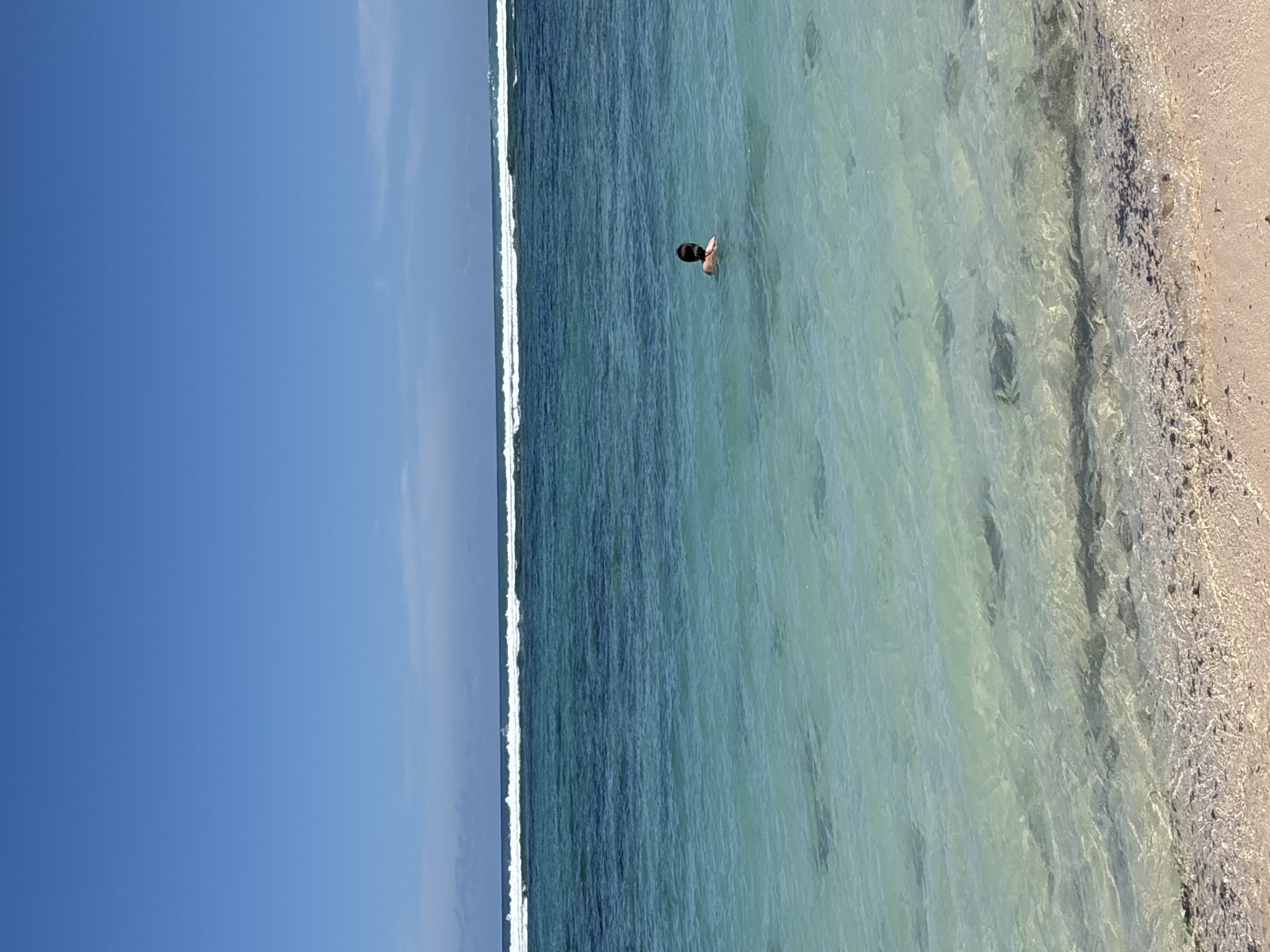 The image depicts a beach scene in Kuta Selatan, Indonesia. The sky is mostly clear with a gradient from a lighter haze to a deeper blue. The ocean stretches across the center of the frame with waves breaking near the shore. A single person is visible swimming in the shallow, clear water. The shoreline consists of a sandy beach.