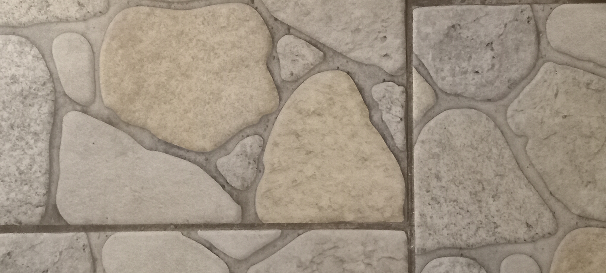 The image shows a close-up, vertical view of a tiled surface with a cobblestone-like pattern. The tiles are irregularly shaped and have a textured, stone-like appearance, with shades of light gray, beige, and cream. The grout between the tiles is a light gray color. There are no people or discernible objects in the image, and the environment is not identifiable beyond the tiled surface. The image does not provide any information that would allow for the localization of this surface within the city of Koviary, Ukraine, or any other specific location.
