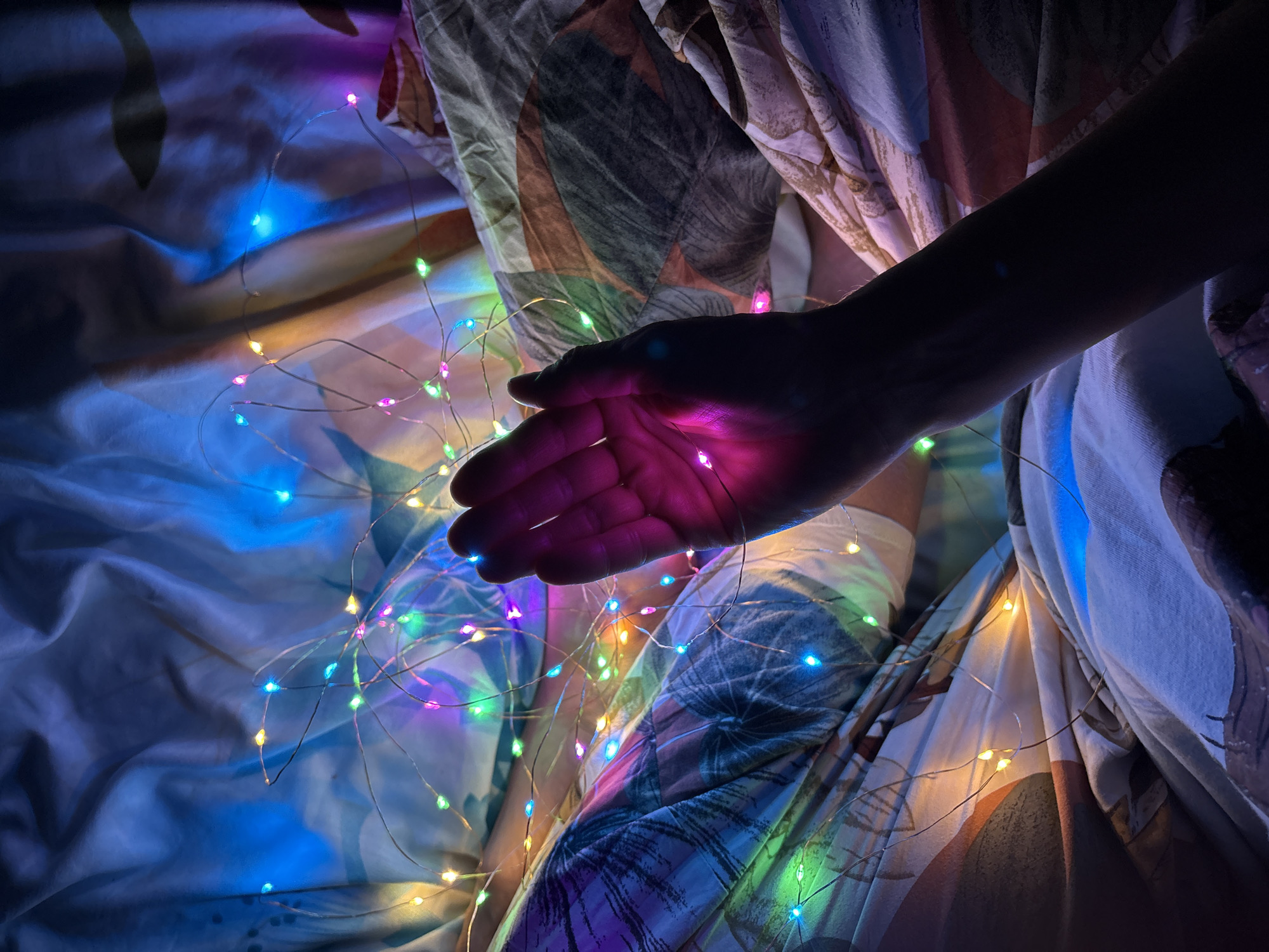 A photograph shows a dark, silhouetted hand held above a bedspread adorned with multicolored fairy lights. The lights illuminate the palm of the hand with a pink hue. The bedspread has a pattern with blue, green, pink and neutral tones. The exact location in the city of Ulyanovsk, Russia is not discernible from the image.
