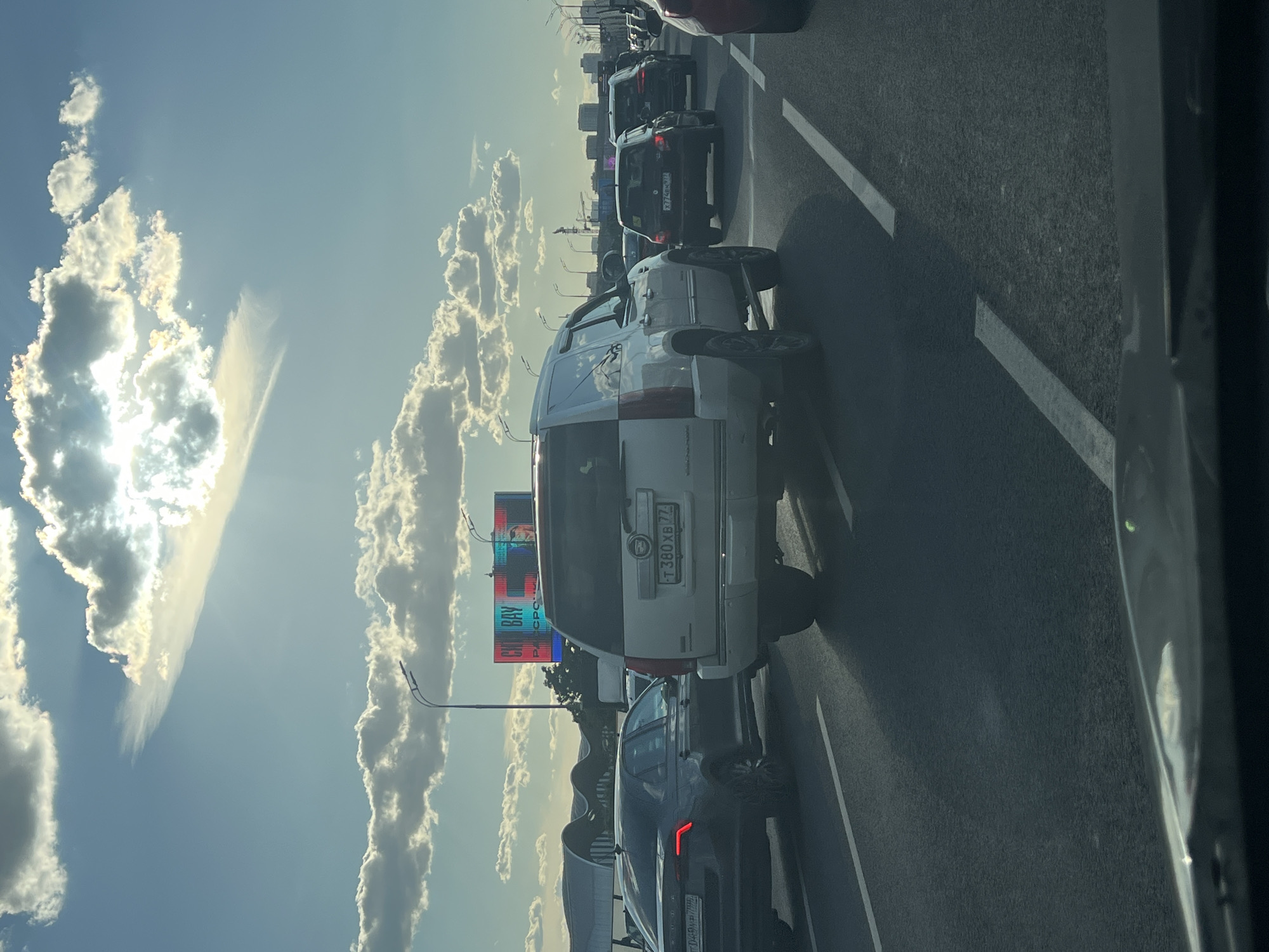 The image shows a congested highway in Moscow, Russia, under a bright, partly cloudy sky. The sun is visible through the clouds, casting rays of light. Several cars are stopped in traffic, including a white Cadillac Escalade in the foreground. The Escalade has a Russian license plate that reads "T380XB 77". Other vehicles visible include a dark gray sedan with its taillights illuminated, a black hatchback, and a black SUV. In the background, there is a large billboard displaying colorful advertisements, and beyond that, a cityscape with buildings and cranes. Streetlights are present along the side of the highway. The perspective suggests the photo was taken from inside another vehicle.