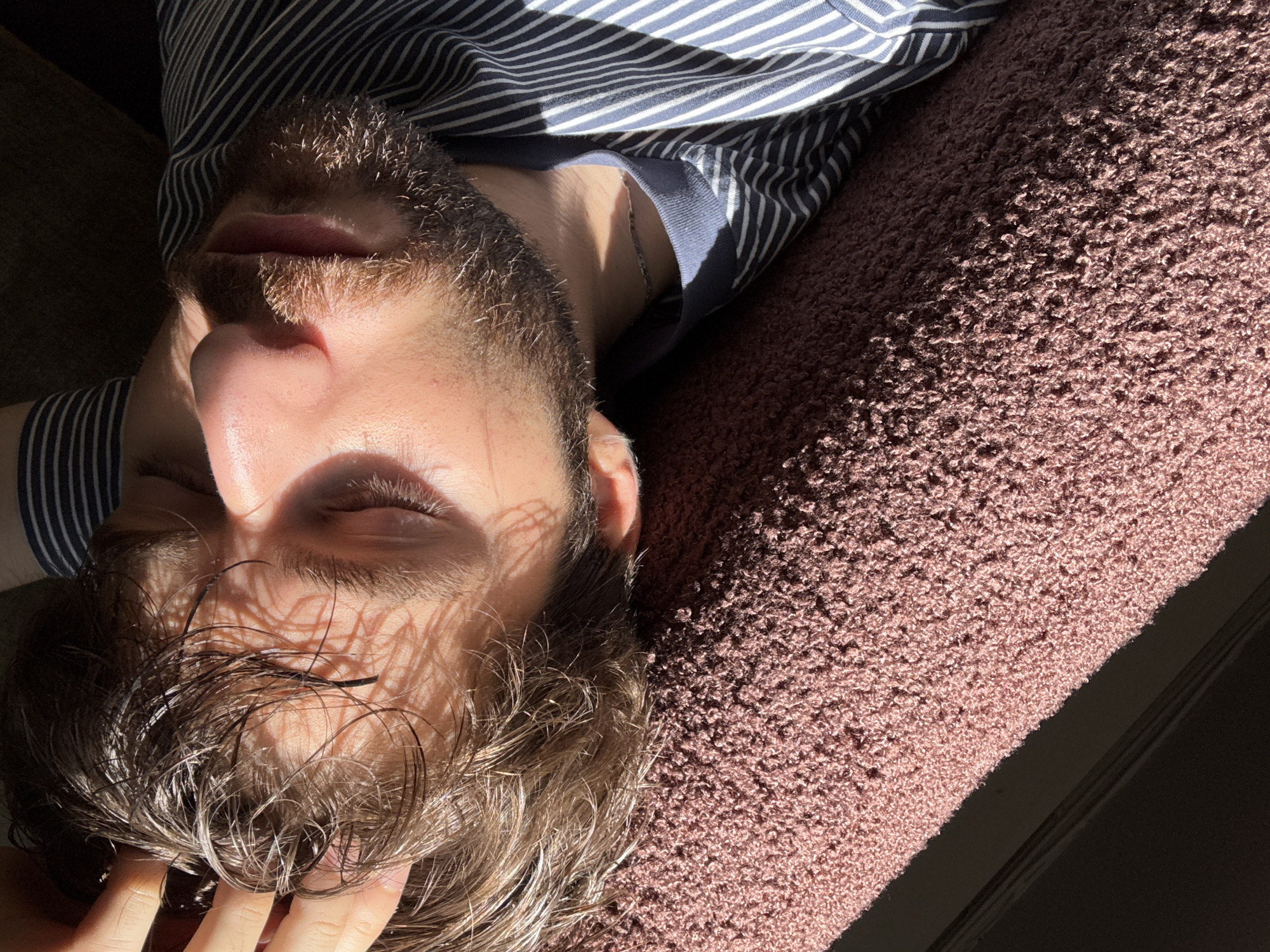 A person with short, wet, light brown hair and a beard is lying on a brown shag rug or blanket. The person is wearing a striped blue and white shirt. Their eyes are closed, and light is streaming across their face, creating shadows from trees. The location is unspecified, but the city is Montreal, Canada.
