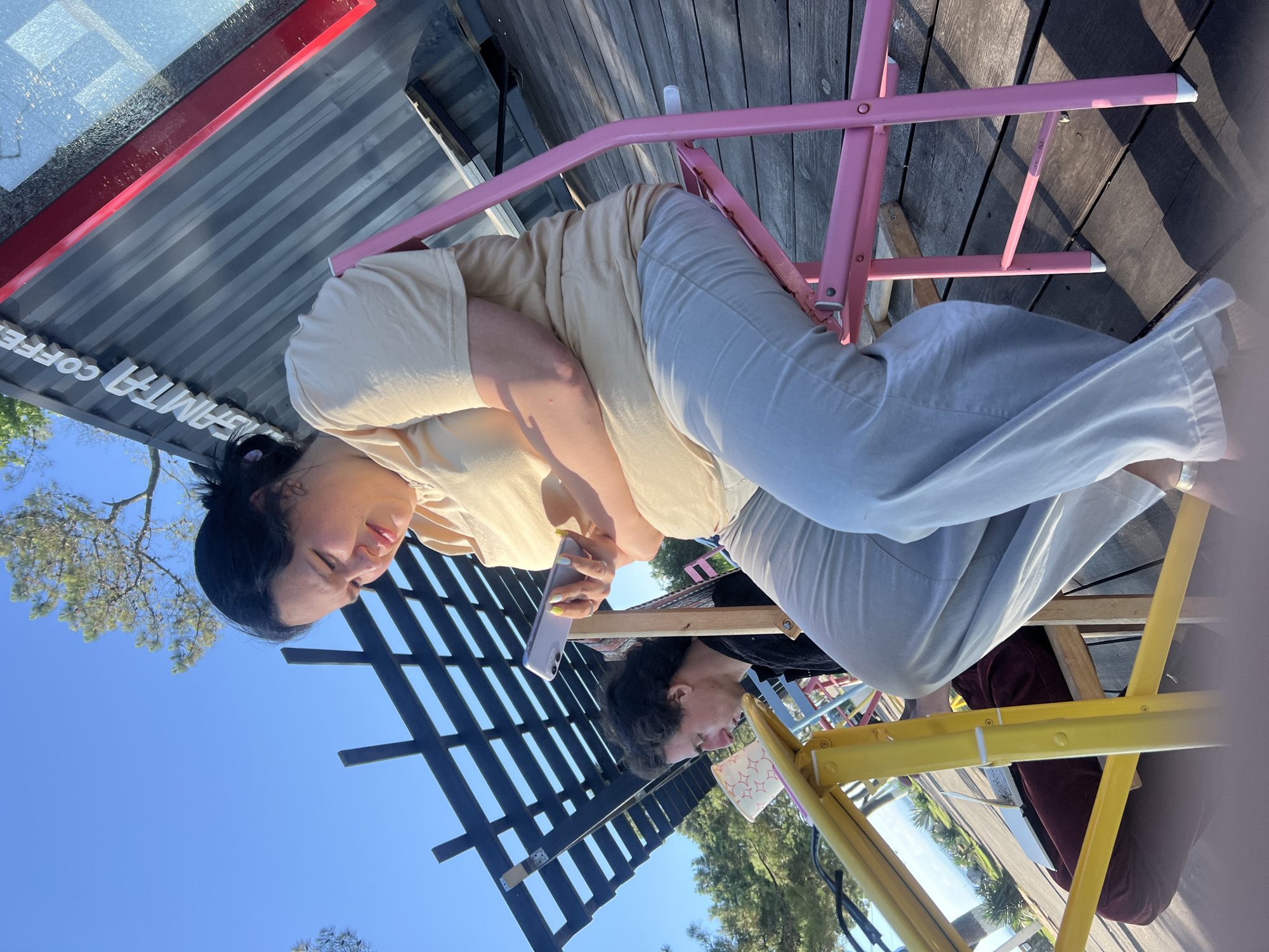 In Ochamchire, Abkhazia, two individuals are seated outdoors. In the foreground, a woman with dark hair pulled back is looking down at her mobile phone. She is wearing a light beige t-shirt and light gray pants and is seated on a pink folding chair. To her left, a man with curly dark hair is looking down at a table. He is wearing a dark shirt. He is seated on a wooden chair. A yellow table is positioned between them, with a white cup with red stars visible on it. The ground is a wooden deck. In the background, a portion of a shipping container structure with white lettering that reads "SAMTA COFFEE" is visible, as is a blue sky and trees.