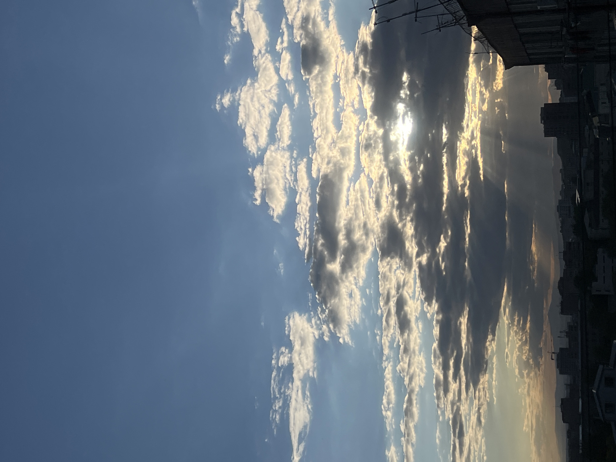 The image shows a view of the sky, partially obscured by buildings at the right edge. The sky is mostly blue on the left, transitioning to a mix of white, gray, and dark clouds toward the right. The sun is visible through the clouds, casting rays downward. The buildings appear to be located in a city, with rooftop details and silhouettes of structures visible. It is impossible to confirm that it is in the city of Yerevan, Armenia. There are no people visible.