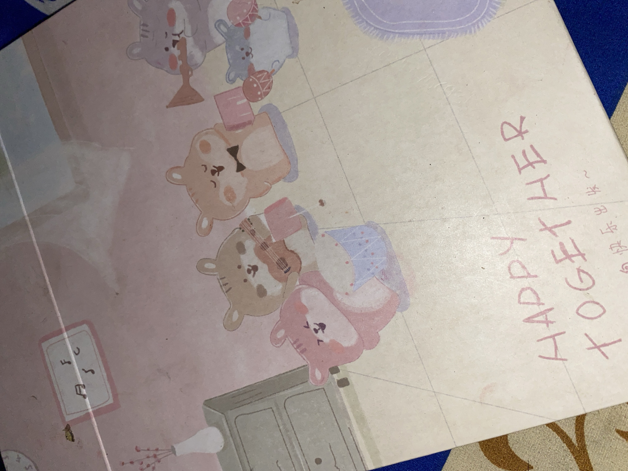 The image displays a close-up of a notebook or sticker sheet with a pastel pink background. The artwork features several cartoonish hamster characters. In the upper left corner, a framed area contains musical notes. A vase with pink floral sprigs sits on a grey cabinet to the left.
The central part of the illustration shows a group of hamsters. One hamster, in light brown, plays a ukulele. Another, also light brown with a black bowtie, stands with a pink rectangular object. A pink hamster is seated and appears to be playing a drum. To the right, a grey hamster plays a trumpet, and a smaller blue hamster holds maracas.
The lower portion of the image has handwritten text in pink. The visible English text reads "HAPPY TOGETHER." Below this, there is some Chinese text. The overall aesthetic is cute and whimsical.
The image is taken from an angled perspective, with the bottom right corner showing a blue and beige patterned fabric. There is no discernible information within the image that would indicate its location in Ta', Indonesia.