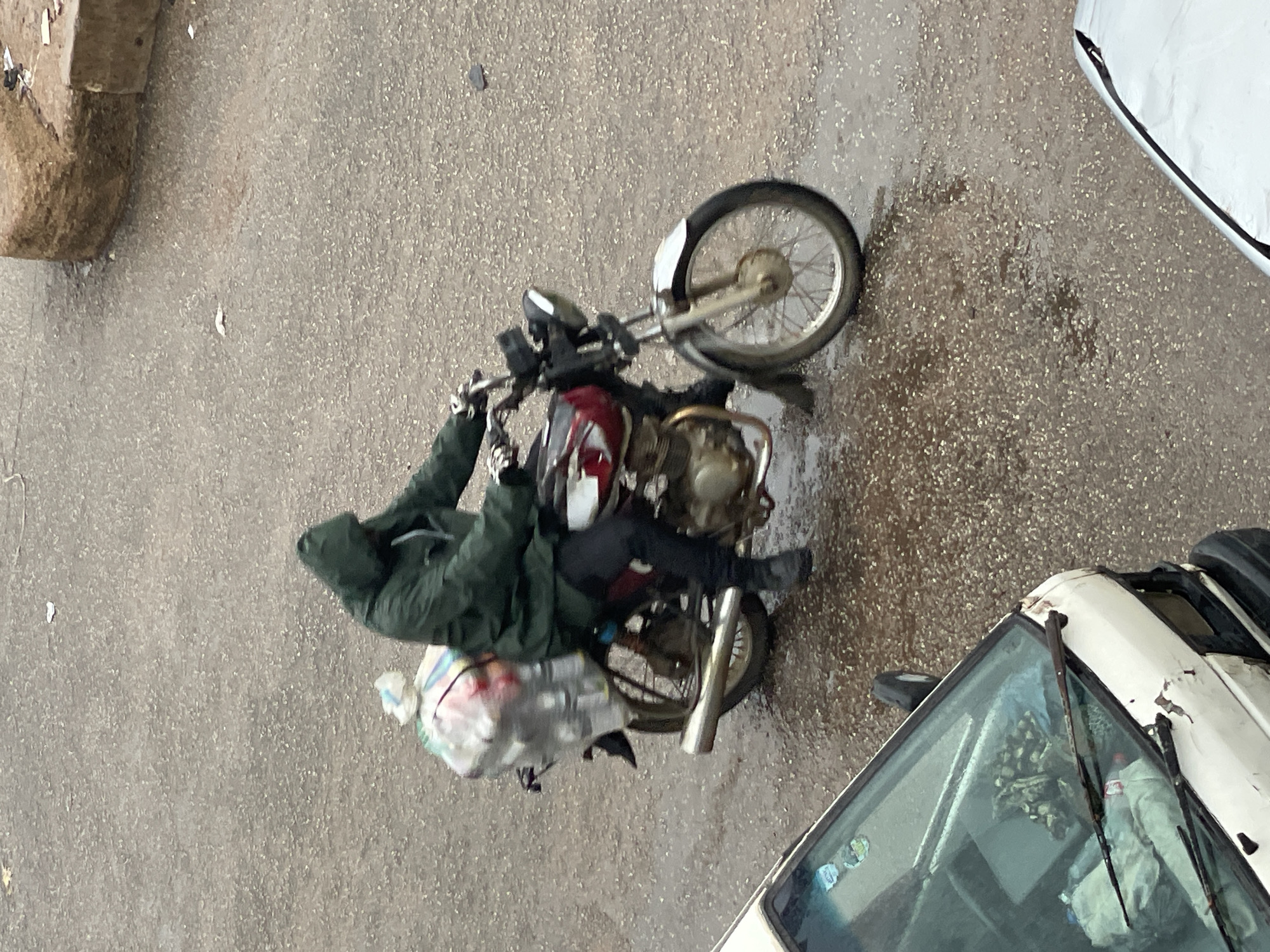 A person wearing a dark green hooded jacket rides a red and white motorcycle on a wet, grey asphalt surface. The motorcycle is carrying a large, clear plastic bag filled with items strapped to the back. The person's face is obscured by the hood. The environment appears to be outdoors in a city, with parts of two white vehicles visible in the foreground and to the left. The ground is wet, indicating recent rain or damp conditions.