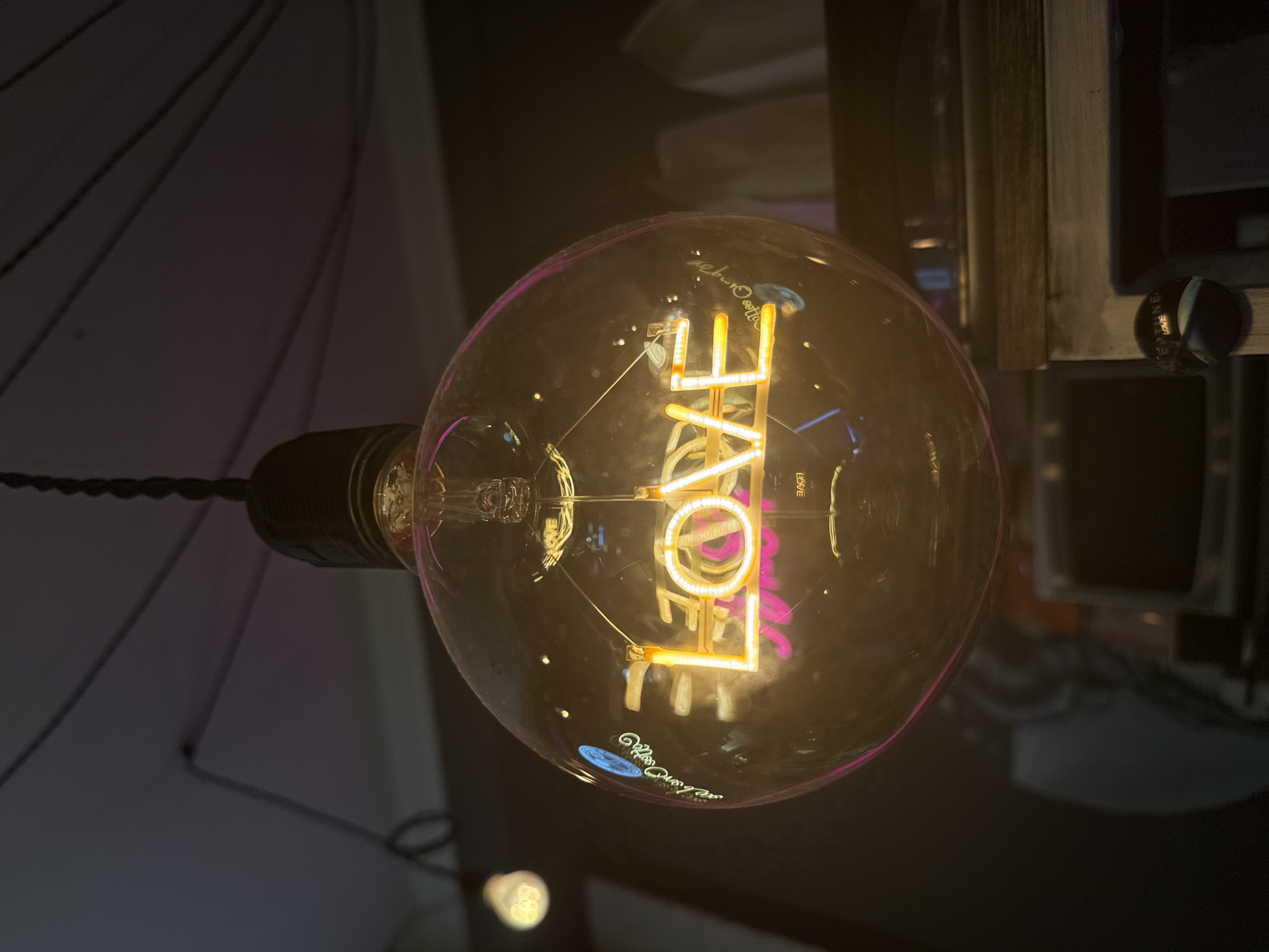 A close-up, eye-level shot focuses on a large, round incandescent lightbulb suspended from a black cord. Inside the glass bulb, a neon sign spells out the word "LOVE" in bright yellow capital letters, outlined with pink. The filament of the bulb is visible behind the neon sign. Reflections on the bulb's surface show parts of the surrounding environment, including a dimmer light source in the background on the left and what appears to be wooden furniture or shelving on the right. The overall lighting is dim, with the neon "LOVE" sign being the primary light source within the bulb. The image does not contain any discernible elements that would allow for a specific location in Moscow, Russia, to be identified.