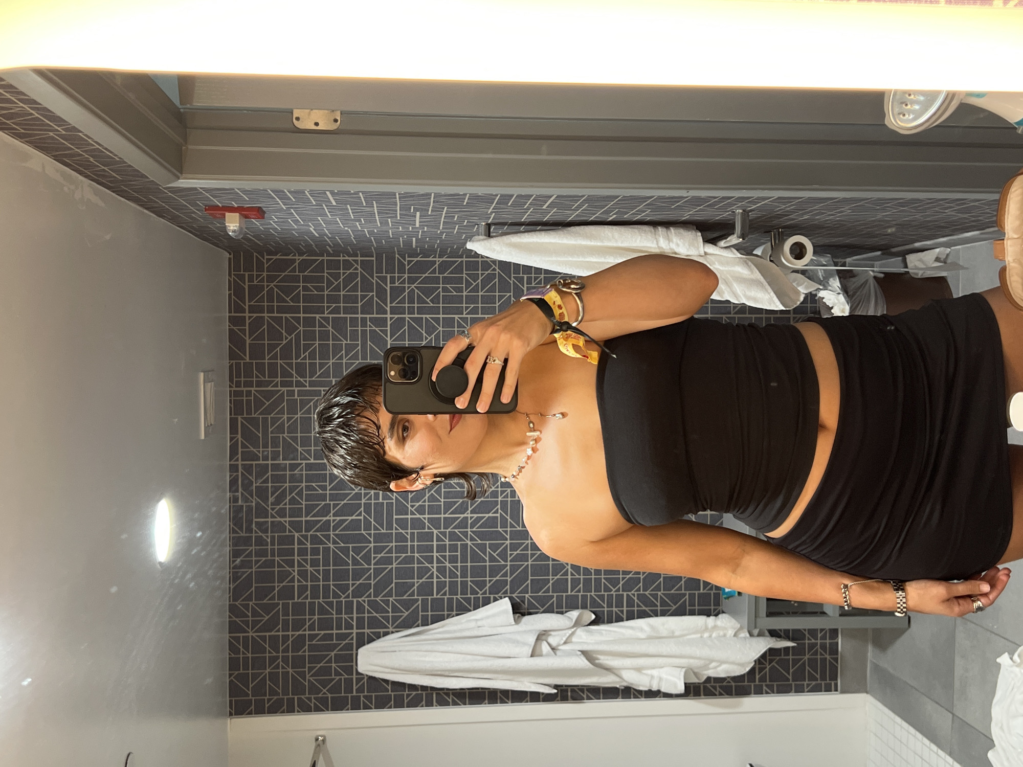A woman is taking a selfie in a bathroom. She is wearing a black tube top and skirt. The bathroom has a modern design with gray and white tiles and a geometric pattern on the wall. A white towel and a few bathroom amenities are visible. Based on the prompt, the location is La Quinta, United States of America.
