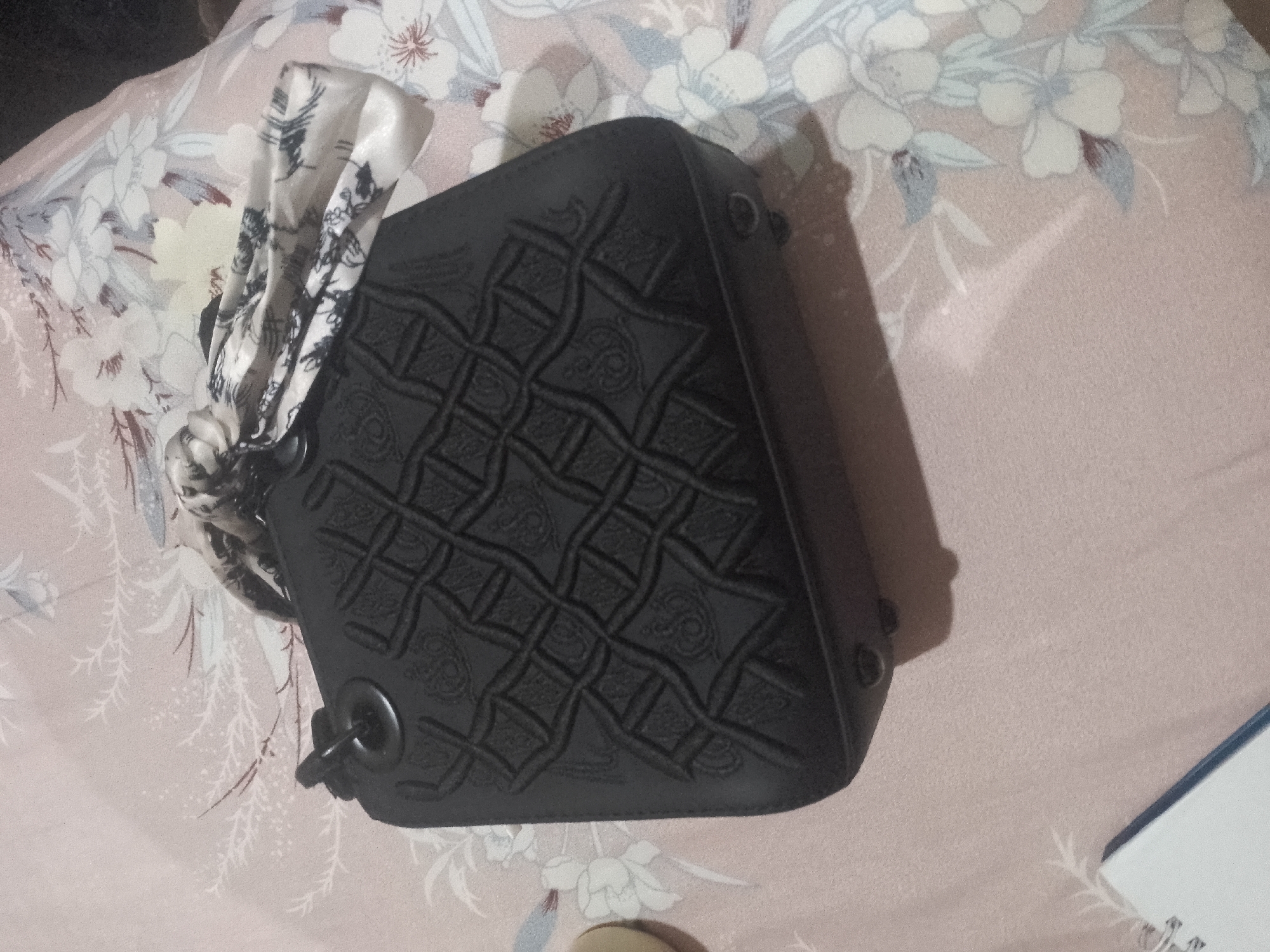 A black leather handbag with a decorative embossed pattern sits on a pink and white floral patterned bedspread. A light-colored scarf with a black and white pattern is tied to the bag's strap hardware. The scene is illuminated from the left, casting a shadow of the bag onto the bedspread. A corner of a blue and white spiral-bound notebook is visible in the lower right of the frame.