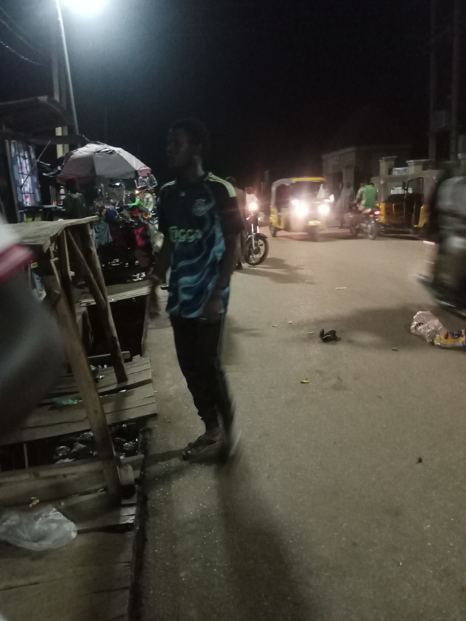 The image depicts a street scene in Minna, Nigeria, at night. A man wearing a blue and black t-shirt with the "Cicco" logo is walking on the sidewalk beside a wooden structure, possibly a market stall. The road is paved and shows signs of wear, with scattered debris including a discarded sandal, plastic bags, and food wrappers.
In the background, several motorcycles and auto-rickshaws (locally known as "Keke Napep") are visible, their headlights illuminating the street. There are some people on and around the motorcycles. There are also covered vendors in the background. The lighting is provided by a street lamp and the headlights of the vehicles. The overall atmosphere suggests a busy street market or gathering place at night.