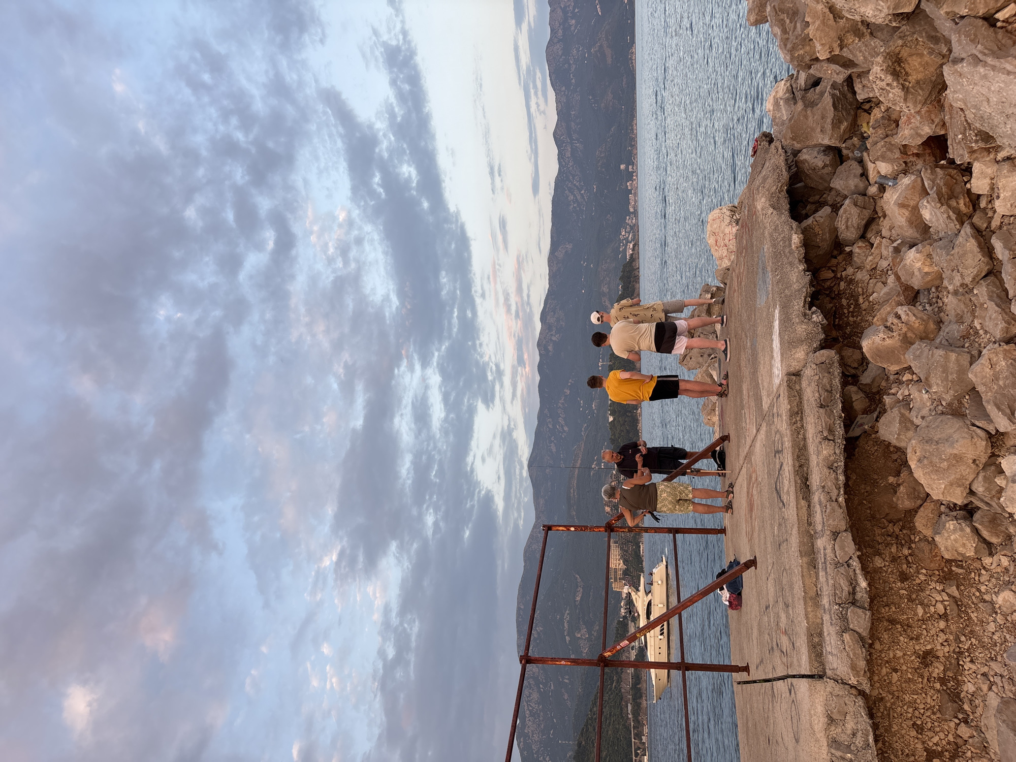 The image depicts a group of four people on a damaged concrete pier overlooking the sea in Budva, Montenegro. A large white yacht is docked in the water to the left, with a town and mountainous landscape in the background. The sky is filled with dramatic clouds tinged with pink and orange, suggesting either sunrise or sunset. The pier itself is broken and eroded on the right side, with large rocks scattered around its base.
Of the four individuals, three are younger men, and one appears to be an older woman. One of the younger men, wearing a yellow t-shirt and black shorts, stands facing away from the camera. Next to him, another young man in a light-colored t-shirt and shorts faces the same direction. A third young man, wearing a white cap and patterned t-shirt, stands beside them. The older woman, wearing a green top and patterned shorts, is facing towards the sea and appears to be speaking with one of the men. The group seems to be engaged in conversation or observing the surroundings. In the foreground, there are piles of rocks and debris, indicating the pier's disrepair. A rusty metal railing runs along the edge of the pier.