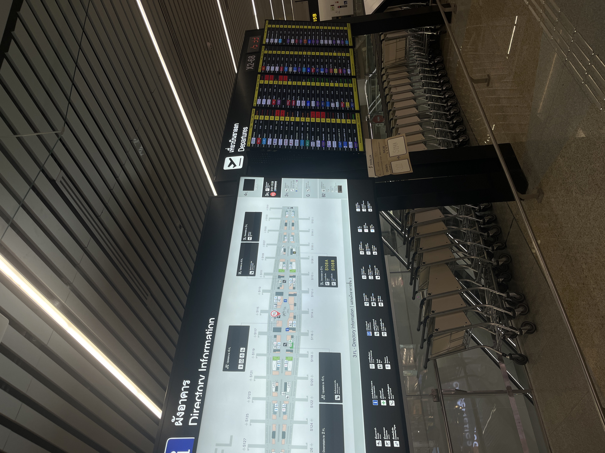 An interior shot captures a well-lit airport environment in Nong Prue Subdistrict, Thailand. The scene is dominated by two large digital display boards, one on the left showing "Directory Information" with a map of airport gates and amenities, and another on the right displaying "Departures" with flight information. The departure board shows flights scheduled for various destinations, including destinations like Hong Kong, Singapore, and Kuala Lumpur, with some flights marked as delayed or cancelled. Adjacent to the departure board, a smaller display shows the gate number X2-68. In the foreground, a row of empty luggage carts are neatly parked. The ceiling features modern linear lighting. The overall impression is one of an organized and functional airport terminal.