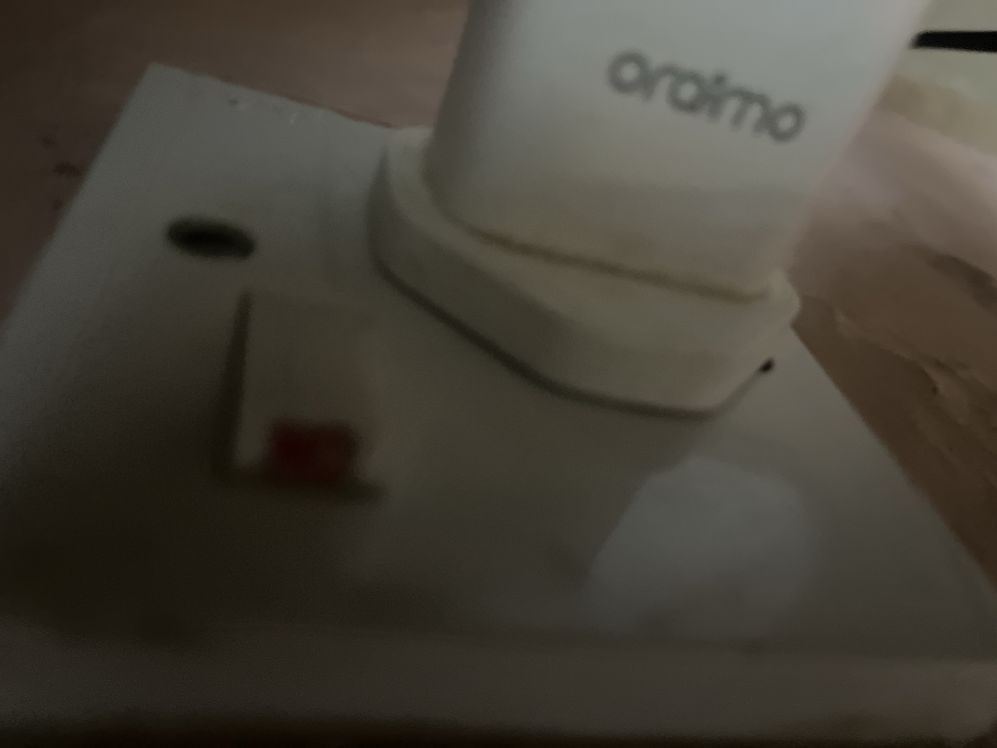 A white electrical outlet is mounted on a wall. A white charger is plugged into the outlet. The charger has the logo "Oraimo" on it. The wall appears to be painted a light pink color. There is a small red button on the outlet. The image is out of focus and taken at a close-up angle.
