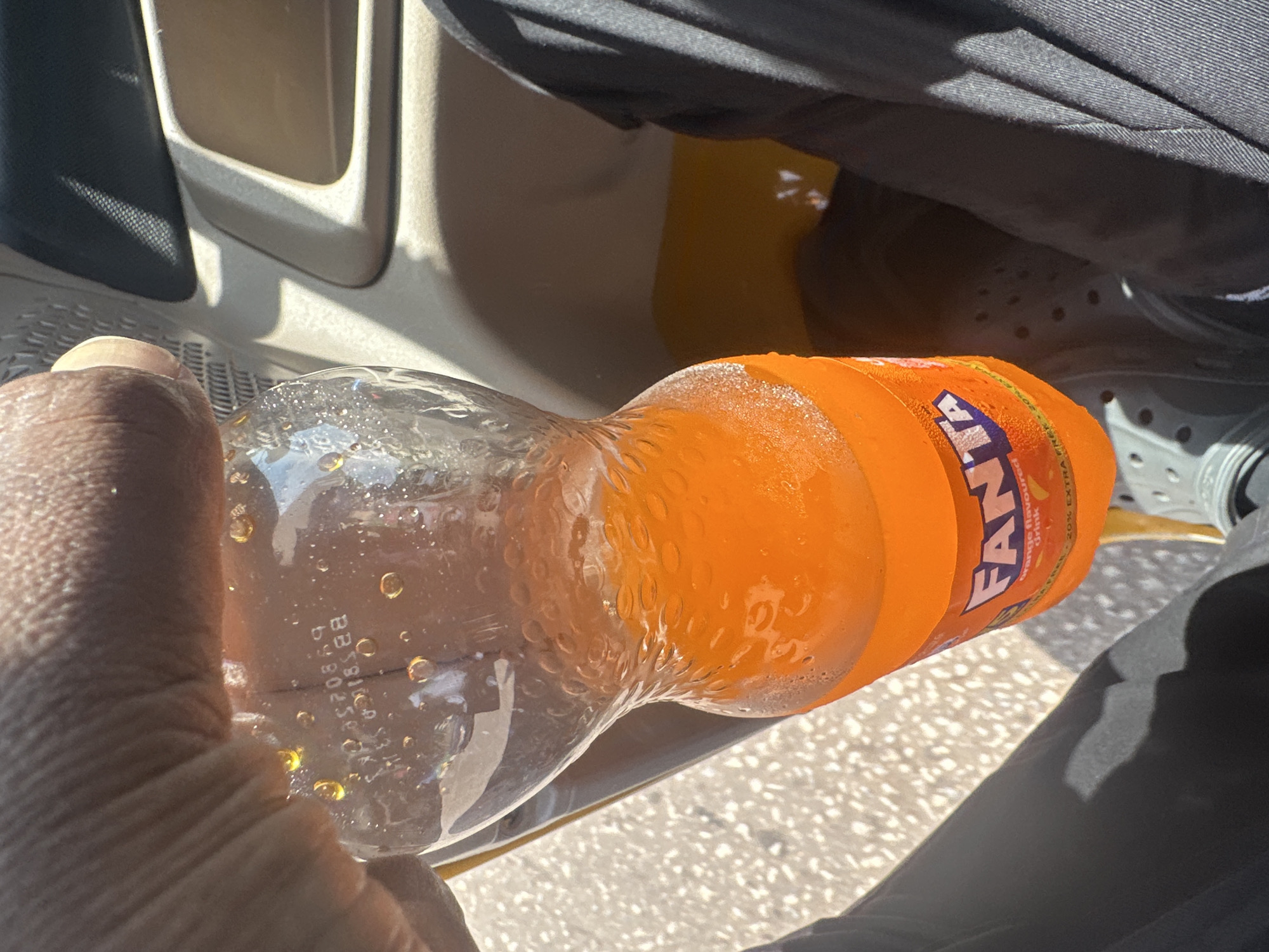 A hand, likely belonging to a person, grips a clear plastic bottle filled with an orange, bubbly liquid. The bottle is partially obscured by the person's fingers and thumb, and condensation droplets are visible on the exterior, suggesting it is cold. The liquid inside also has numerous small bubbles. The bottle has an orange label with white and blue lettering that reads "FANTA". Below "FANTA", it says "Orange flavoured drink". To the right of this, "20% EXTRA" is visible in smaller text.
The bottle is placed within the interior of a vehicle, as indicated by the beige and black plastic trim of a car door or console visible in the upper left of the image. The floor of the vehicle appears to be textured, possibly a rubber mat. Shadows suggest it is daytime, and the lighting indicates bright sunlight. The scene appears to be outdoors, likely during the day, with hints of a sunny environment. The overall impression is casual, possibly depicting someone taking a drink during a journey or break.