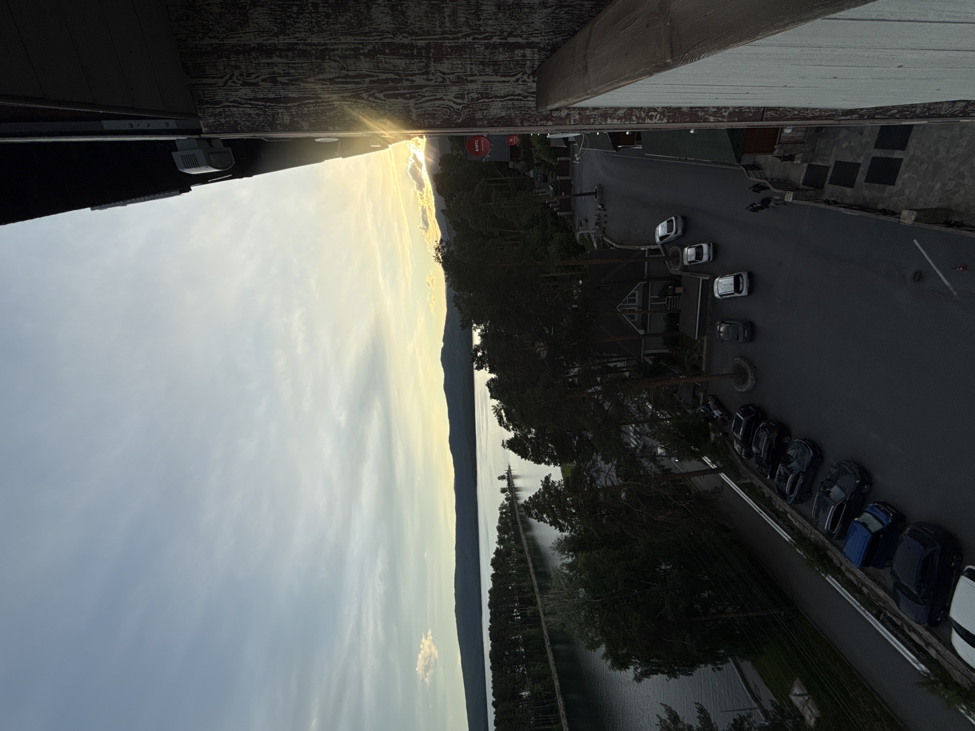 The image shows a view from an elevated position, possibly a balcony, in Dom Otdykha Zolotoy Plyazh, Russia. A body of water stretches into the distance, reflecting the sky. Trees line the shoreline and a road runs parallel to the trees, with several parked cars visible. On the other side of the road are various buildings. In the distance, the sun is setting, creating a bright glow. The sky is filled with clouds. The balcony or building structure is visible at the top and bottom of the frame.