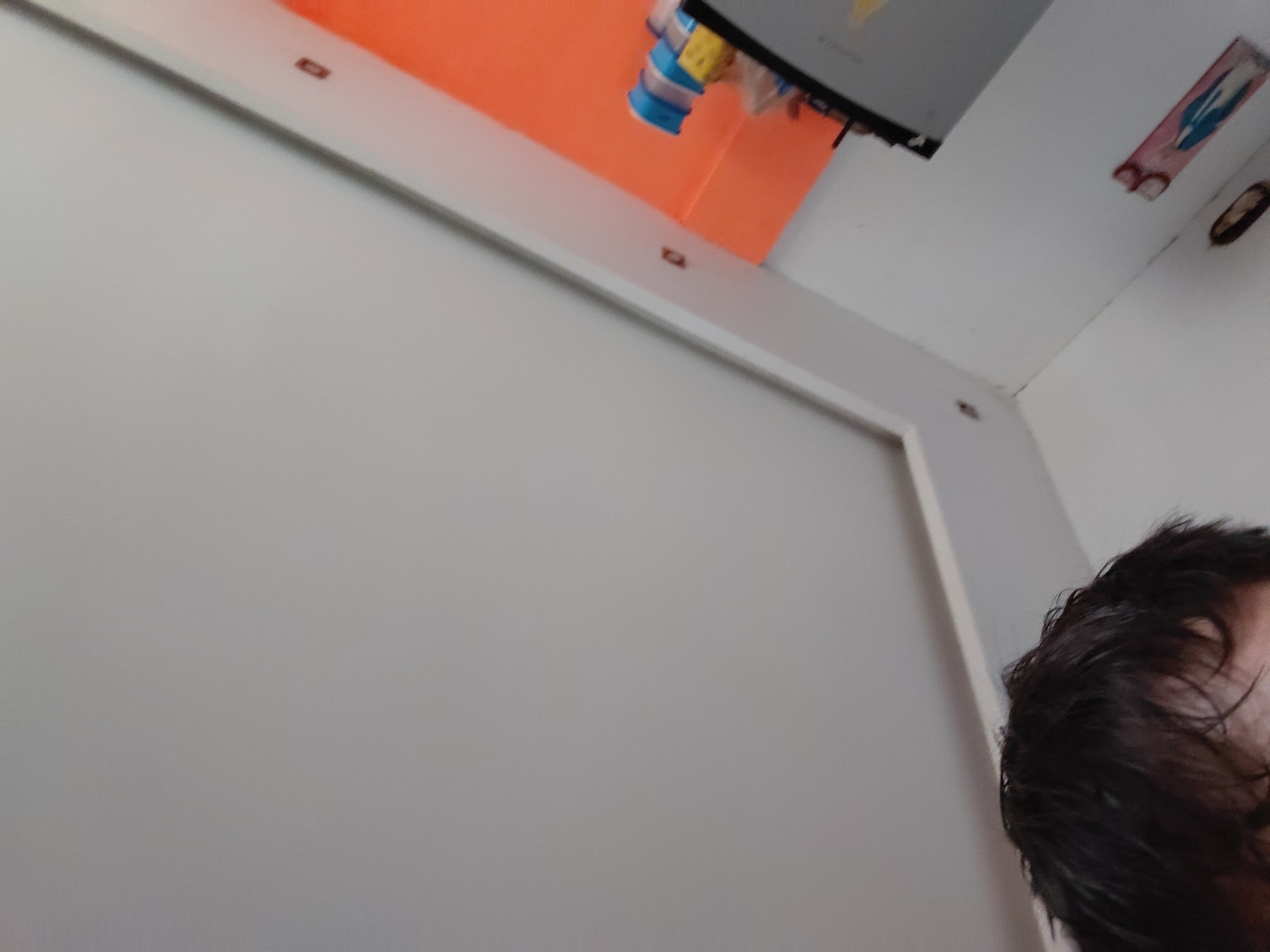 This indoor photograph, taken in Santa Ana, Venezuela, captures a skewed perspective of a room. In the bottom right corner, a partial view of a person's head with dark hair is visible, suggesting the image may be a selfie or taken from a low angle. The majority of the frame is dominated by a light grey wall or ceiling area, bordered by a distinct white frame or beam. Above this white frame, a bright orange surface extends, likely a wall or a large decorative panel.
In the upper central area, a black object, possibly a television screen or monitor, is visible, with small, colorful items (blue and yellow) hanging below it, resembling clips or small toys. Further to the right, on a white wall surface, there is a small, rectangular sticker or picture featuring a blue graphic on a pink background. Part of a red character, which appears to be a "3" or a "B," is visible on this sticker. Below this, a partially obscured oval-shaped object, possibly a light fixture, can be seen. Small, dark, rectangular elements are spaced along the white frame, potentially fasteners or decorative details. No specific activity is discernible beyond the act of photography, and the time of day is not indicated.
