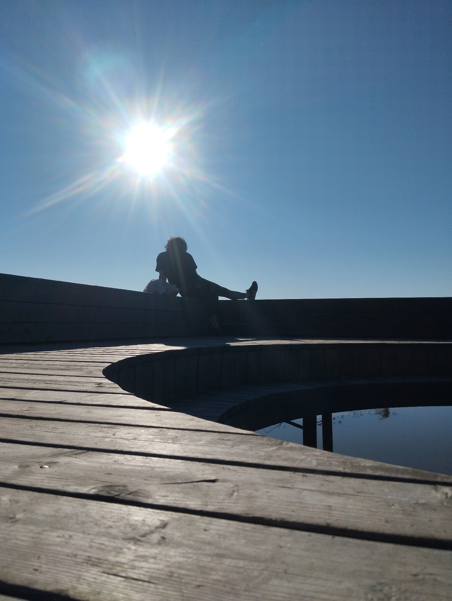 A silhouetted person sits on a wooden platform or deck, seemingly near a body of water. The sun is positioned high in the clear sky, causing significant lens flare. The scene is dominated by a wooden structure with a curved edge, suggesting a pier or observation point. Reflections are visible on the water's surface. The location is Unknown, Unknown.