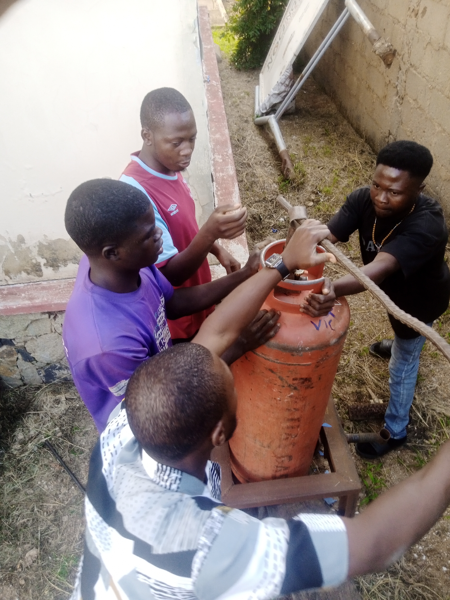 Four young men are gathered around an orange propane tank in an outdoor setting. The propane tank is on a metal stand. The men are wearing casual clothing. One man in a black t-shirt and blue jeans is holding a metal rod that is connected to the propane tank. Another man in a purple t-shirt is also touching the propane tank. Two other men are standing near the tank. The background shows a wall made of concrete blocks and dry grass. There is also a white sign mounted on a metal frame behind the men. The scene appears to be in an urban or semi-urban environment, possibly a yard or an alleyway.