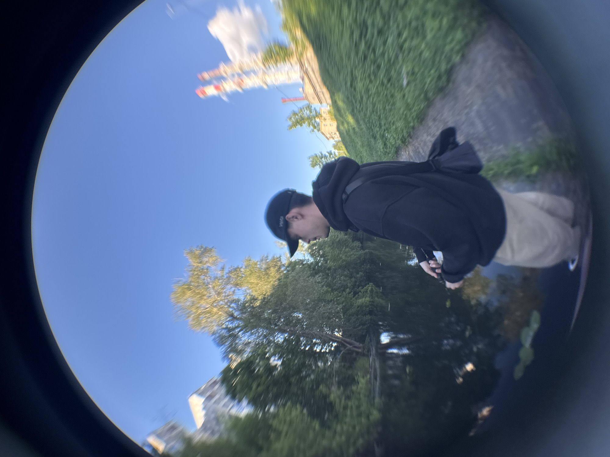 A person wearing a black hoodie and a black baseball cap is standing on a paved path next to a body of water. The person is holding a fishing rod and appears to be fishing. The water reflects the surrounding trees and sky. In the background, there are trees, buildings, and industrial chimneys. The scene is captured with a fisheye lens, creating a distorted, wide-angle view.