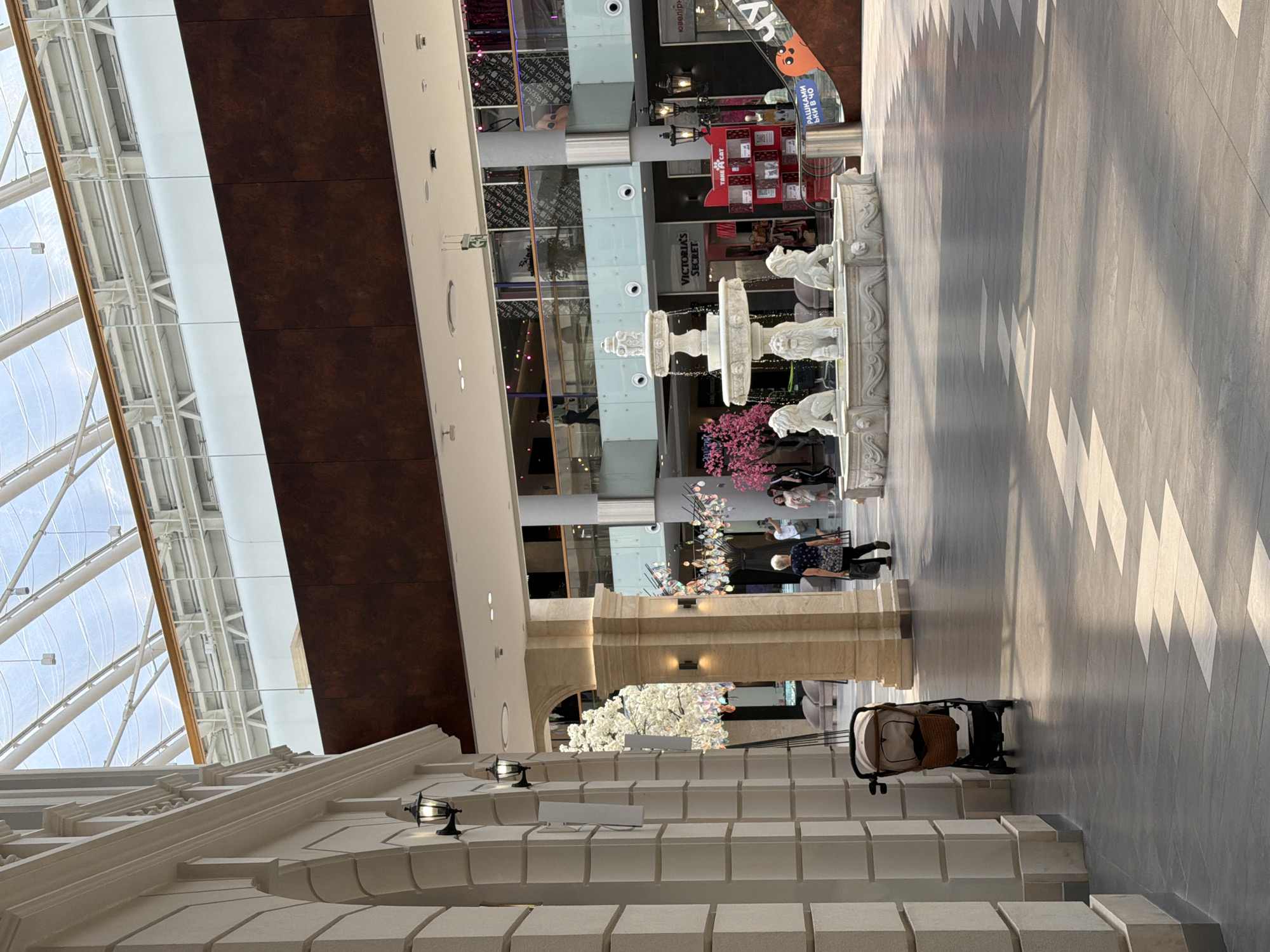 The image depicts the interior of a modern shopping mall, likely in Kyiv, Ukraine, characterized by its spacious atrium and contemporary architectural design. Sunlight streams in from a large, multi-faceted glass ceiling, illuminating the polished stone floor. On the left, a cream-colored architectural facade with arched details and mounted black lanterns lines the walkway. A single baby stroller, a light beige and brown model, stands unattended near the facade.
Dominating the central area is an ornate, multi-tiered stone fountain featuring lion sculptures at its base. Beyond the fountain, several shops are visible, including a "Victoria's Secret" store. People are scattered throughout the mall; a woman in a dark top and patterned skirt walks away from the camera towards the fountain, and other individuals are seen in the background, browsing or walking. Decorative trees with pink blossoms add to the ambiance. The overall environment suggests a well-maintained and active public space, consistent with a high-end shopping center.