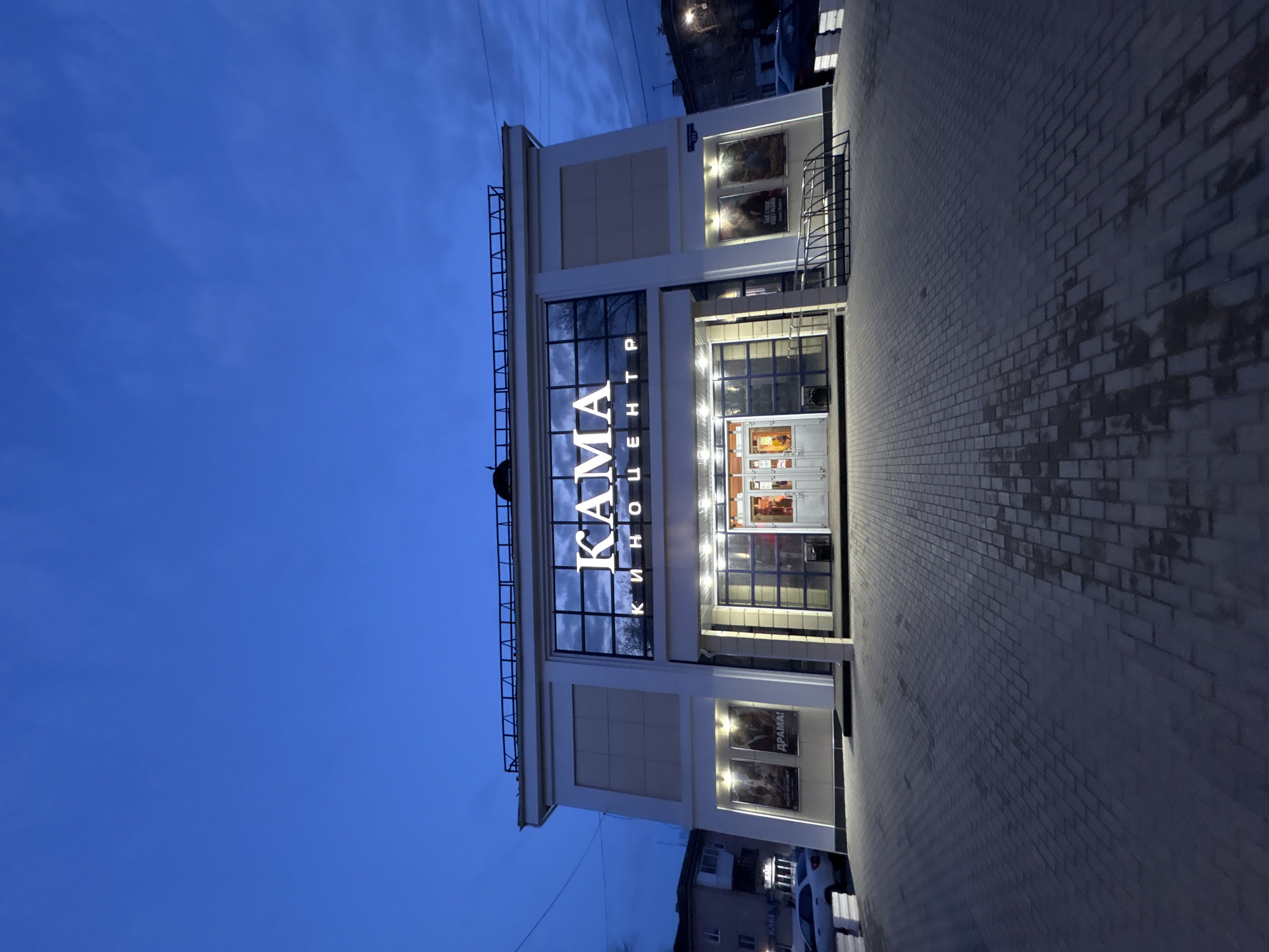 The image depicts the "Kama Cinema Center" located in Chaikovsky, Russia at dusk. The building is a two-story structure with a modern facade featuring large windows and a prominent illuminated sign displaying "KAMA" in Cyrillic script. The entrance is well-lit, with multiple doors visible. Movie posters are displayed on the lower and upper portions of the building's exterior. The ground is paved with light-colored bricks, and the sky is a deep blue, indicating the time of day. There are cars parked nearby and other buildings can be seen in the background.