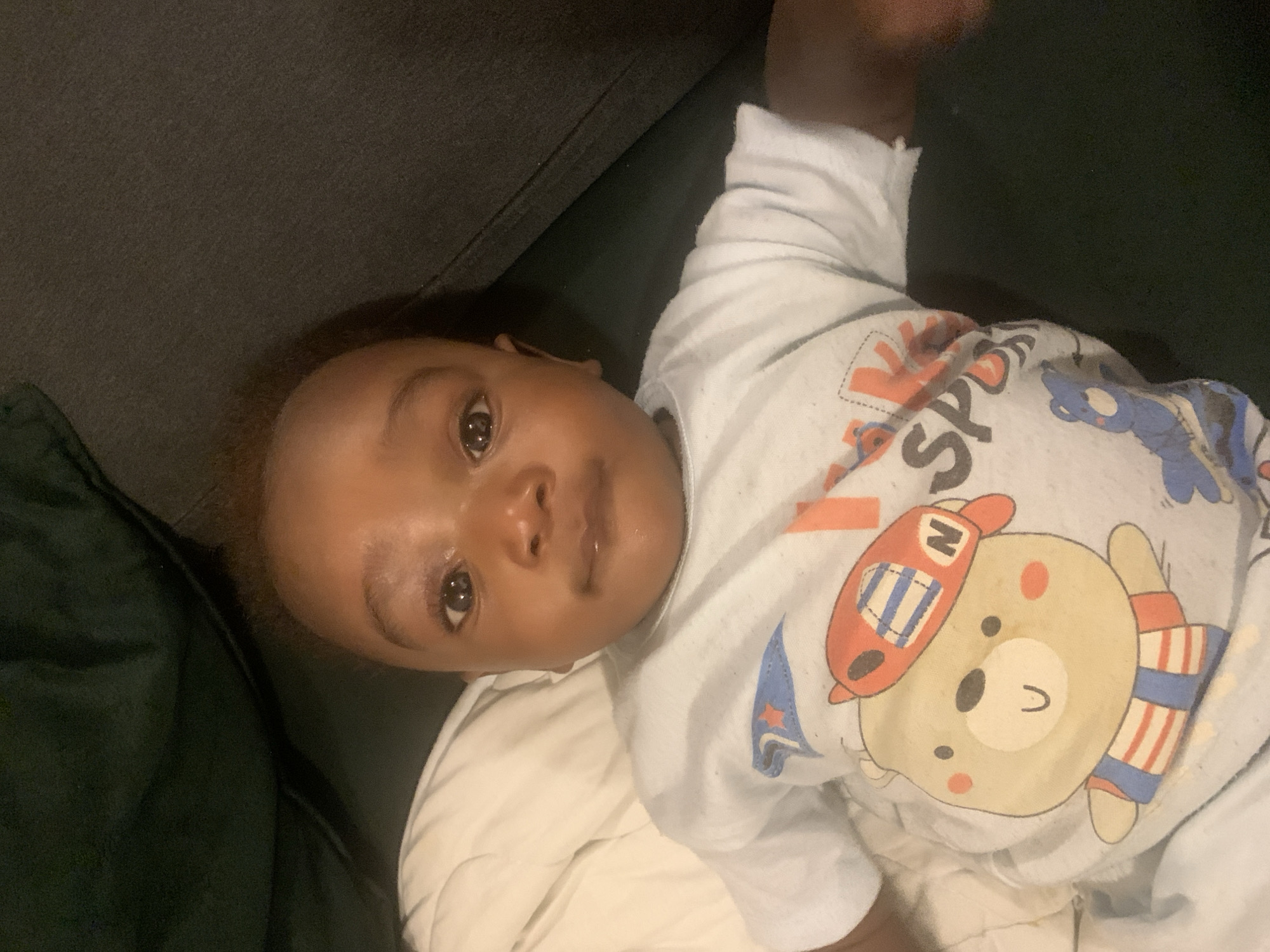 A baby is lying on a dark grey sofa. The baby is dark skinned, wearing a light blue and white shirt with a cartoon bear design. A white blanket or cushion is beneath the baby. The location is unknown, and there is no information to confirm the location in Barakin Akawo, Nigeria.