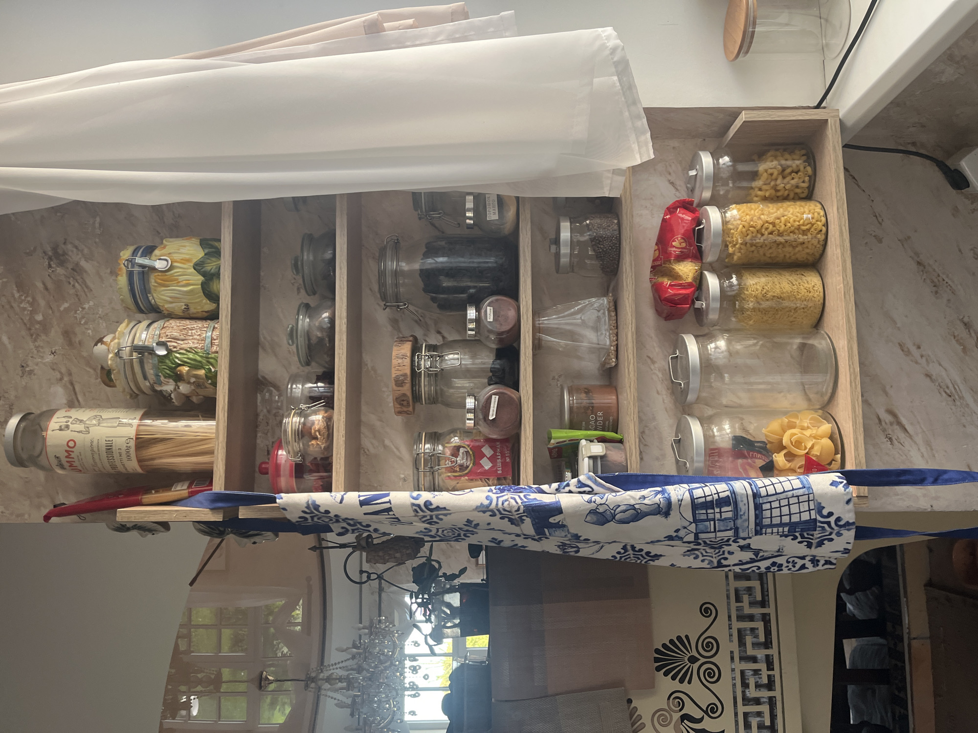 The image displays a wooden shelf unit filled with various food items in jars and containers. On the top shelf, there's a tall jar of spaghetti, a ceramic jar with a mushroom design, and another ceramic jar with a sunflower design. Below that, several glass jars hold dried goods and spices, including what appears to be whole peppercorns, dried mushrooms, and other granular substances. A blue and white patterned apron is hung on the side of the shelf unit. The shelf unit is positioned against a wall with a textured finish. In the foreground, a countertop is visible with more glass jars containing pasta, including macaroni and smaller shaped pasta. A red bag of pasta is also on the shelf. The overall scene suggests a kitchen or pantry setting. The image does not provide any information to suggest the location is Vladimirovka, Russia.