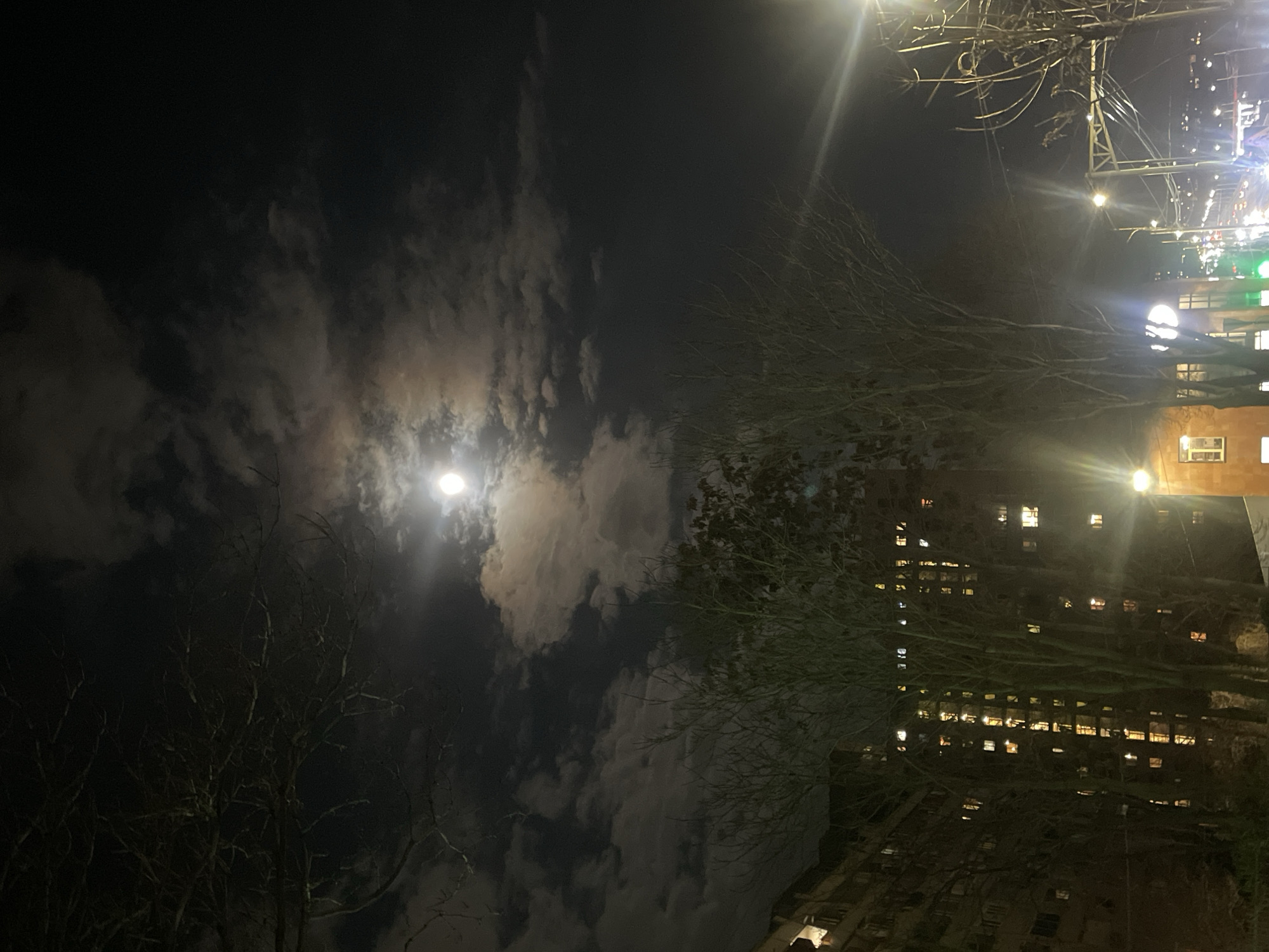 The image depicts a night scene, likely in an urban setting, under a sky partially obscured by clouds with a bright moon visible. Bare tree branches are silhouetted against the sky, and a building with numerous lit windows is visible on the right side of the frame. Artificial light sources, possibly streetlights, create glare and illuminate the scene.
Given the likely Yerevan location, it could be a view of a residential or commercial building.