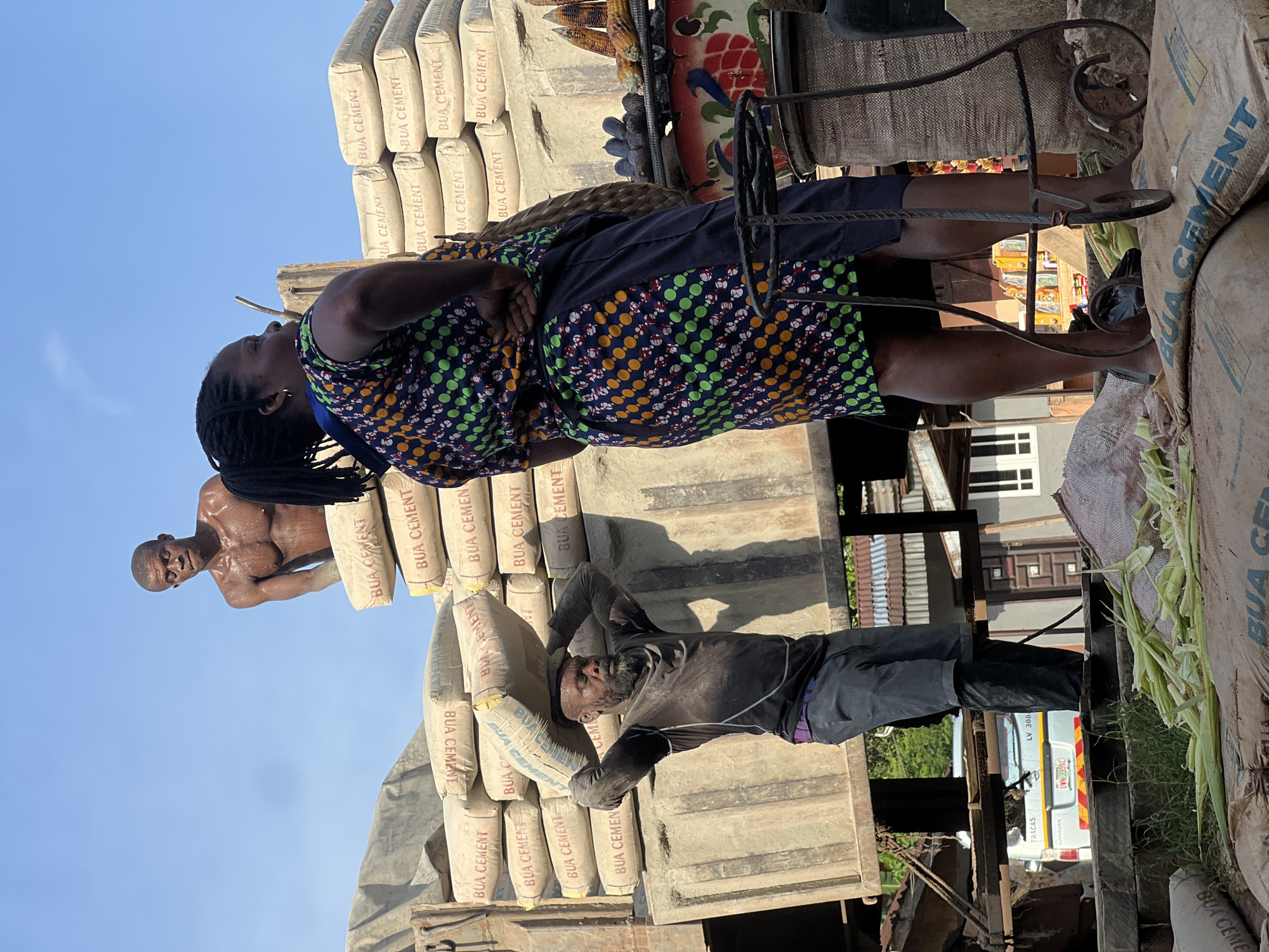 In Nkpor, Nigeria, at a bustling market scene, two men are engaged in the strenuous task of unloading sacks of cement from a large truck. The man on the left, with a rugged beard and sweat glistening on his bare chest, is carrying a heavy sack of cement on his head. His muscular physique is evident as he balances the load. Another man, shirtless and equally drenched in sweat, is visible behind him, seemingly having just handed off a sack or preparing to receive one. In the foreground, a woman stands with her back to the camera, her colorful patterned dress and dark skirt creating a vibrant contrast against the dusty, utilitarian backdrop of the market. She appears to be observing the activity or perhaps waiting for her turn. The truck is piled high with more sacks of cement, all bearing the "BUA CEMENT" logo. Piles of corn husks and some cobs of corn are scattered around, suggesting the proximity of other market goods. The environment is bright and sunny, with a clear blue sky overhead. The overall impression is one of hard work and commerce in a lively urban setting.