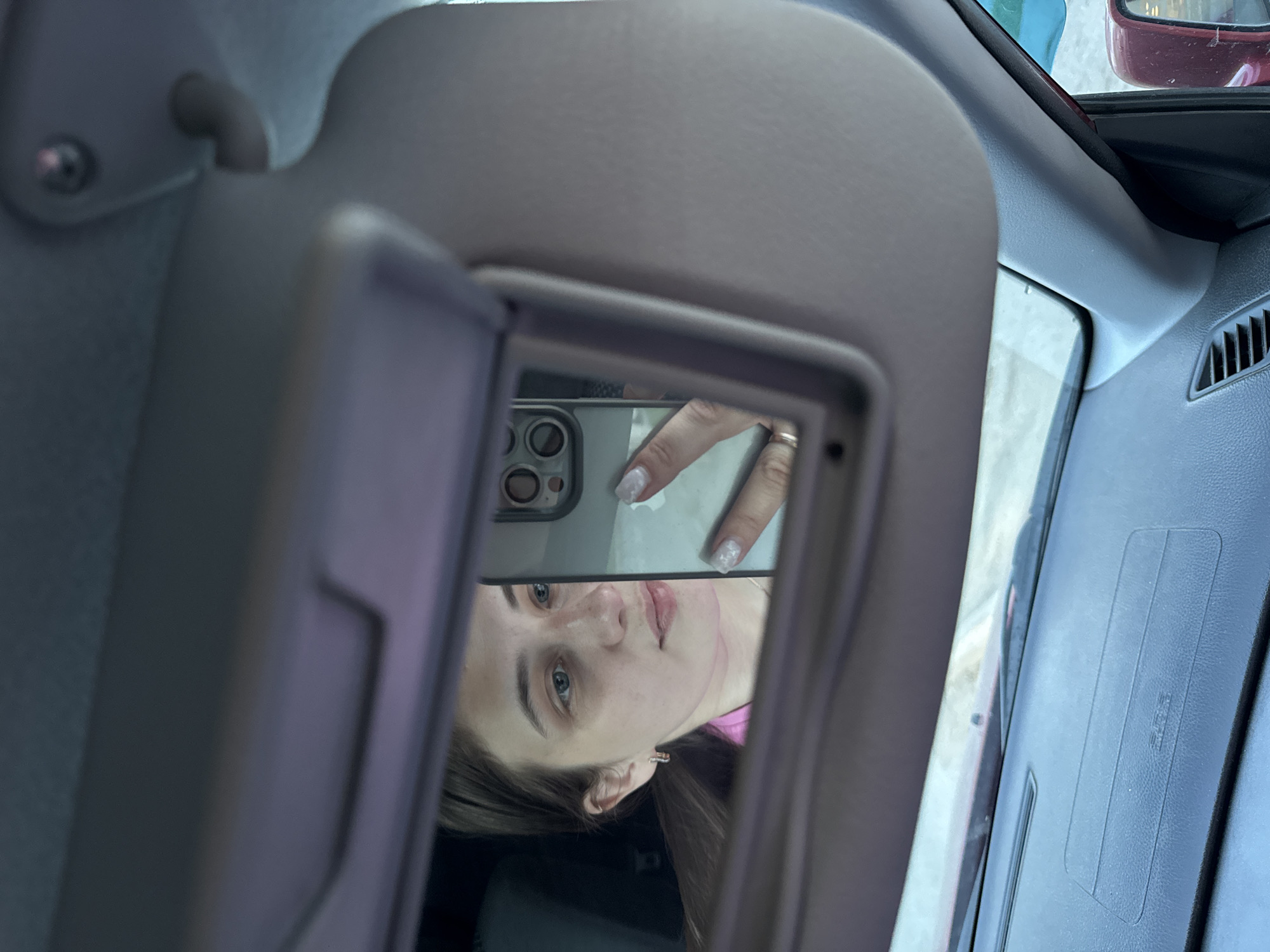 The image shows a selfie being taken in the rearview mirror of a car. The photographer's face and hand holding the phone are reflected in the mirror. The car's interior is visible around the mirror, including the sun visor. Outside the car, there are glimpses of other vehicles, partially obscured. The city of Chaikovsky, Russia is not evident in the image.