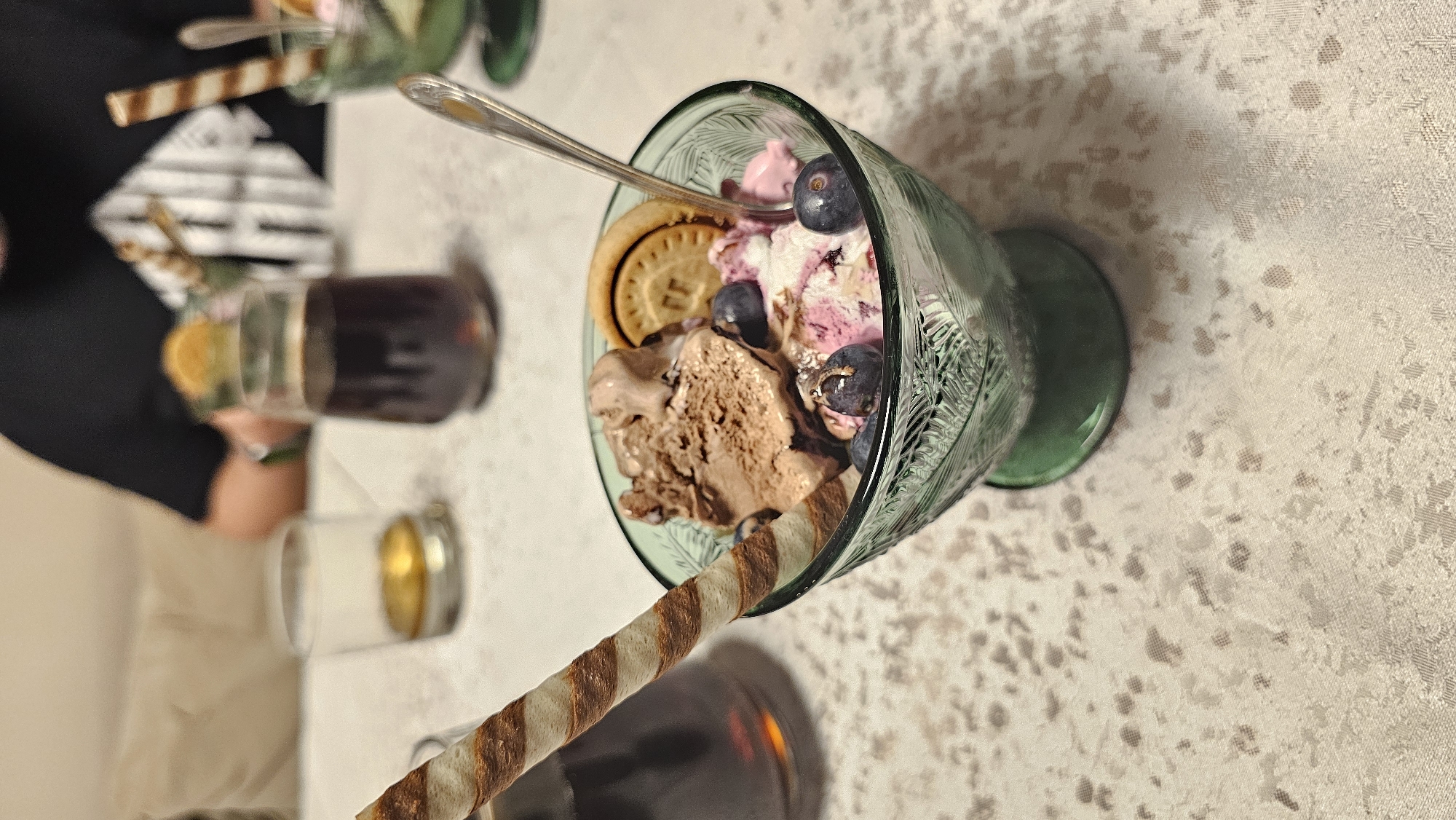 A top-down, close-up shot presents a glass filled with ice cream and toppings. The glass, green with a textured design, contains a scoop of chocolate ice cream, a scoop of pink and white ice cream, blueberries, and a round biscuit. A silver spoon is partially submerged within the ice cream. A striped wafer stick leans against the glass. In the blurred background are other similar glasses of dessert, a tall drink with an orange slice, and a person seated at the table wearing a black shirt with a geometric design. The table is covered with a patterned beige tablecloth. The location is likely a restaurant or cafe, possibly in Varna, Bulgaria, but this cannot be conclusively confirmed from the image.