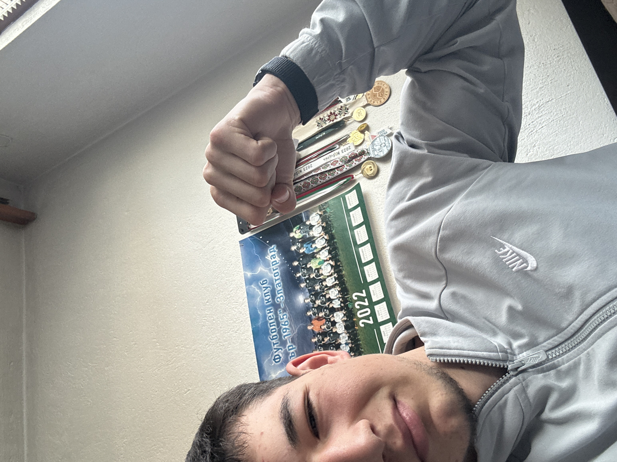 A young man takes a selfie, capturing his upper body and right arm, against a plain light-colored wall. He is seen from the forehead down to just below the chin, with dark hair, eyebrows, and light stubble, sporting a slight, pleasant smile. He wears a light gray zip-up jacket with a visible white "NIKE" logo on the chest.
His right arm is raised, with his hand clenched into a fist. Behind his arm, several medals and awards are displayed on a surface, possibly on a shelf or table. These include ribbons with text such as "2023 KSFULTRA" and "2023 K3", as well as a round, golden, coin-like medal. Behind the man's head and arm, a poster or calendar is visible, featuring a group photo of a football team and the prominent year "2022". Cyrillic text on the poster reads "Футболен клуб" (Football Club) and "Златоград" (Zlatograd), with "ър-1965" likely indicating a founding year or another date related to the club. The overall impression is of a proud individual showcasing his achievements and affiliations, likely within the context of Zlatograd, Bulgaria. The indoor lighting suggests it is daytime.