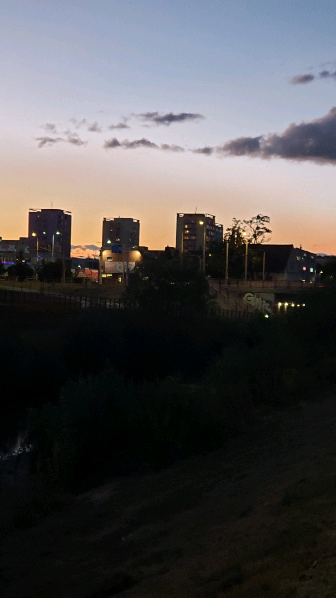 The image depicts a dusk scene in Prešov, Slovakia, featuring the silhouette of residential buildings against a gradient sky of pale blue and orange. Several tall, rectangular buildings with visible windows and lights are prominent in the mid-ground. In the foreground, dark foliage and trees obscure a lower section of the scene, with a hint of a body of water on the far left. A bridge with graffiti is visible in the mid-ground, spanning across a dark area, possibly a river or valley. The overall atmosphere is calm and urban, characteristic of a twilight cityscape. There are no people or animals visible in the frame.