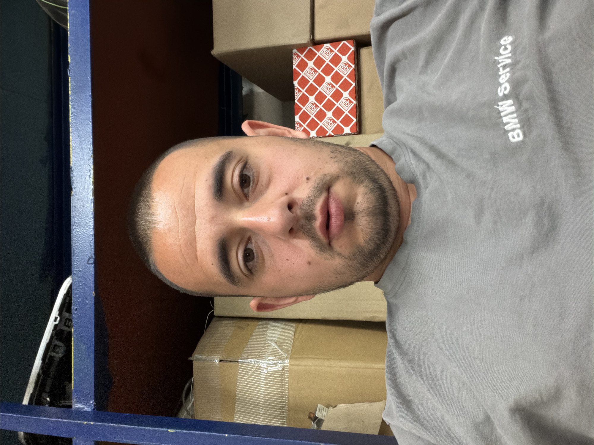 A man with a shaved head and a short beard is pictured in a medium shot, looking directly at the camera. He is wearing a grey t-shirt with "BMW Service" embroidered on it. The background consists of shelves filled with cardboard boxes. The scene appears to be in a storage area, possibly a workshop or warehouse. The lighting is dim, creating shadows on the man's face and the surrounding environment. The image quality is average, with some graininess. The man's expression is neutral. There are no other people or visible actions taking place in the image. The overall impression is one of a casual, informal portrait taken in a utilitarian setting. The image is from Burgas, Bulgaria.