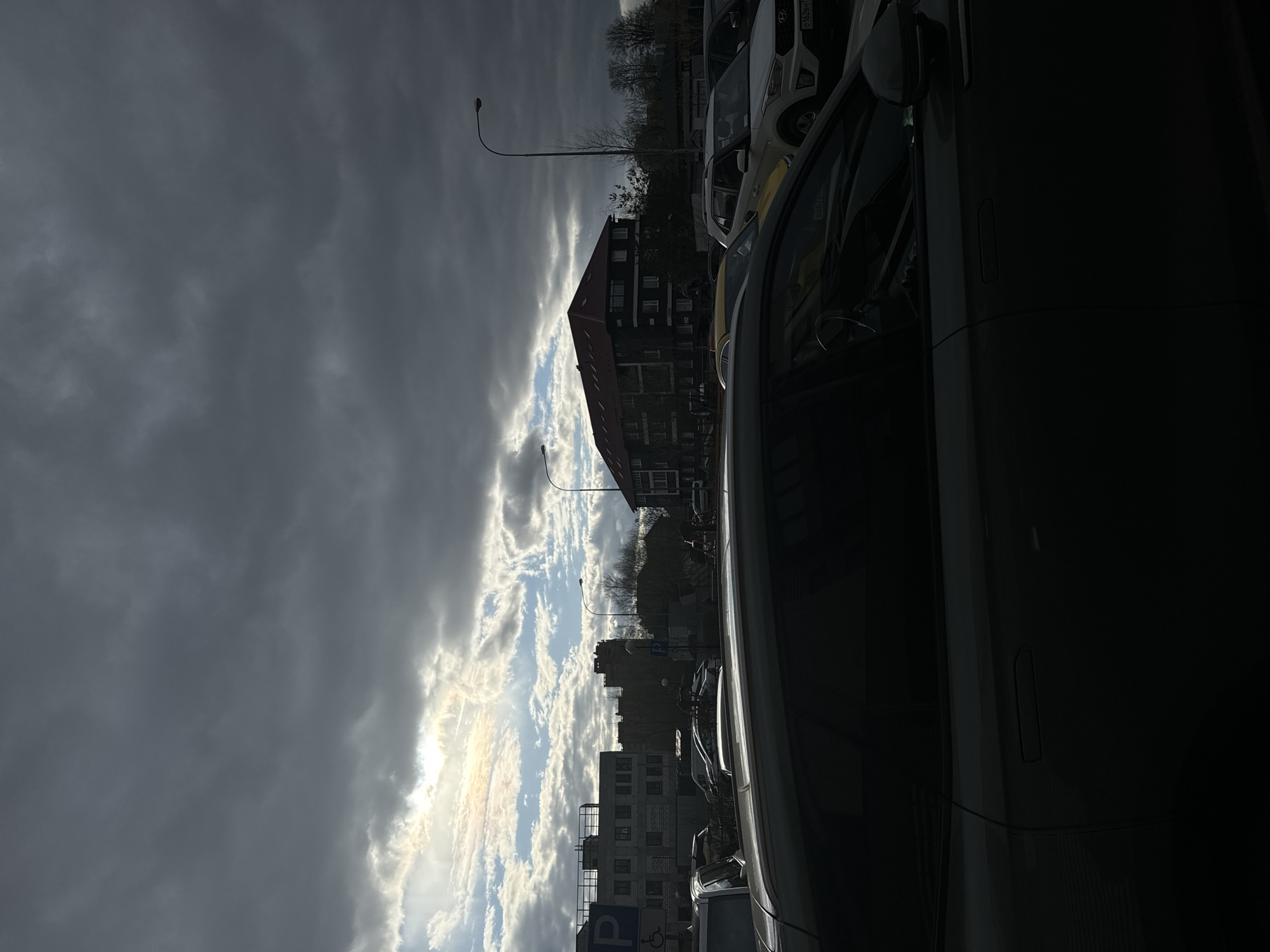 The image, oriented correctly, depicts an outdoor urban or suburban street scene under a dramatic, cloudy sky, likely during late afternoon or early morning. The weather appears overcast with breaks in the clouds, allowing sunlight to dramatically illuminate parts of the sky and creating strong contrast.
In the foreground on the right, the dark side profile of a car is heavily shadowed. Behind it, several other cars are parked, including a white vehicle (possibly a Hyundai Creta) with a partially visible Russian license plate reading "0162BH 7..." and text "3B87B" on its window.
The mid-ground features several buildings: a prominent dark multi-story building with a red roof, and other structures in the distance. Streetlights or lampposts with curved arms rise above the parked cars and buildings. A blue parking sign with a white 'P' and a wheelchair accessibility symbol is visible towards the lower left of the mid-ground. Bare trees are scattered in the background, suggesting a dormant season.
The sky occupies a significant portion of the image, characterized by dark, heavy grey clouds on the left contrasting sharply with bright, sunlit clouds on the right, where the sun appears to be breaking through. The overall lighting is dim due to the extensive cloud cover, but the sun's presence creates striking highlights. No people are visible. The scene is static, showing parked vehicles and architecture. The location is Akulovo (Gp Kubinka), Russia.