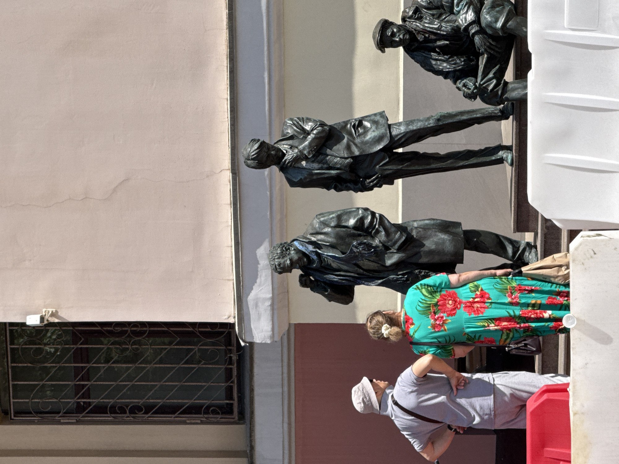 The image depicts a group of people in front of a bronze sculpture in Moscow, Russia. The sculpture appears to be of several figures, some of whom are lying down or in contorted poses, suggesting a dramatic or historical event. One of the figures in the sculpture is a man in a suit, with his head bowed. Another is a man reaching out, seemingly in distress.
Two women and one man are standing in front of the sculpture. One woman is wearing a vibrant green dress with a tropical floral pattern, and a white flower in her hair. She is looking at the sculpture. The second woman is partially obscured by the first. The man is wearing a light-colored cap and a grey t-shirt. He is looking up at the sculpture.
The scene appears to be outdoors, with a building in the background and what looks like a window with a decorative metal grate. The lighting suggests it is daytime, with bright sunlight. The overall atmosphere is one of contemplation or appreciation of the artwork. No text is visible in the image.