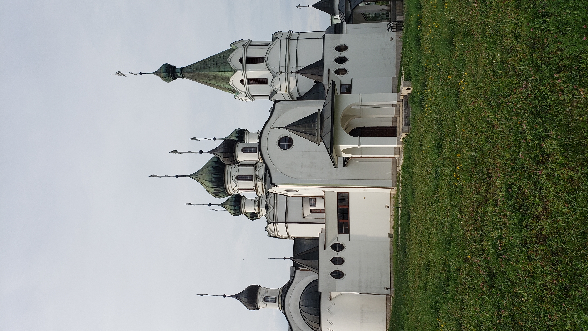 This is a photograph of the Orthodox Church of St. John the Merciful in Prešov, Slovakia. The church is a white, multi-domed structure with green onion domes topped with crosses. The building is set against a pale, overcast sky. In the foreground, there is a lush green lawn dotted with small yellow and pink wildflowers. The church has several entrances, including a covered porch with arched openings leading to a dark wooden door. There are also small windows visible on the facade of the building. The overall scene depicts a peaceful, religious edifice in a natural setting.