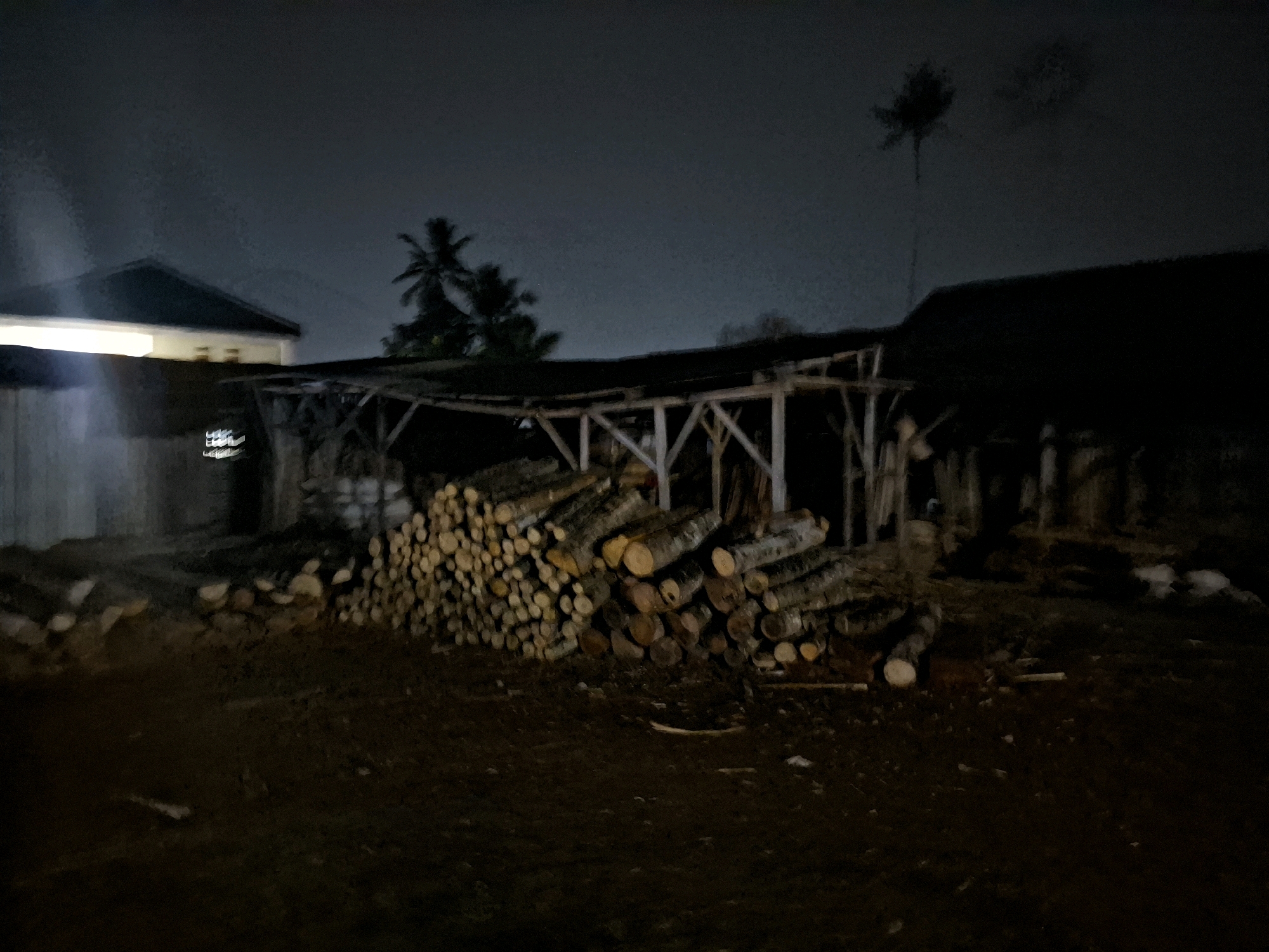 This is a dimly lit, nighttime photograph taken in Ciheulang Tonggoh, Indonesia. The scene depicts an outdoor storage area with a large pile of cut logs stacked under a simple wooden shelter. To the left of the logs is a building with a light shining from an interior space. In the background, there are several palm trees and the dark outlines of more structures. The ground in the foreground is covered with dirt and debris. The overall impression is one of a rural or semi-rural location at night.