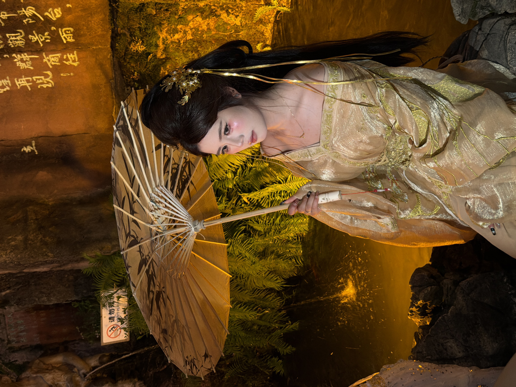 A young Asian woman with dark hair styled in an updo and adorned with a golden floral hairpiece sits in front of a rock face inscribed with Chinese calligraphy. She wears a traditional gold-colored garment with intricate embroidery and holds a partially open, decorative umbrella. Her makeup is prominent, with pink blush on her cheeks and elongated eyeliner. She is seated on rocks beside a body of water where sparks or fire effects are visible. In the background, ferns and other greenery are present, and a sign with a prohibition symbol and Chinese text is affixed to the rock face. The lighting is warm and dramatic, suggesting an evening or indoor setting with artificial illumination. The location is identified as Yuzhong District, China.