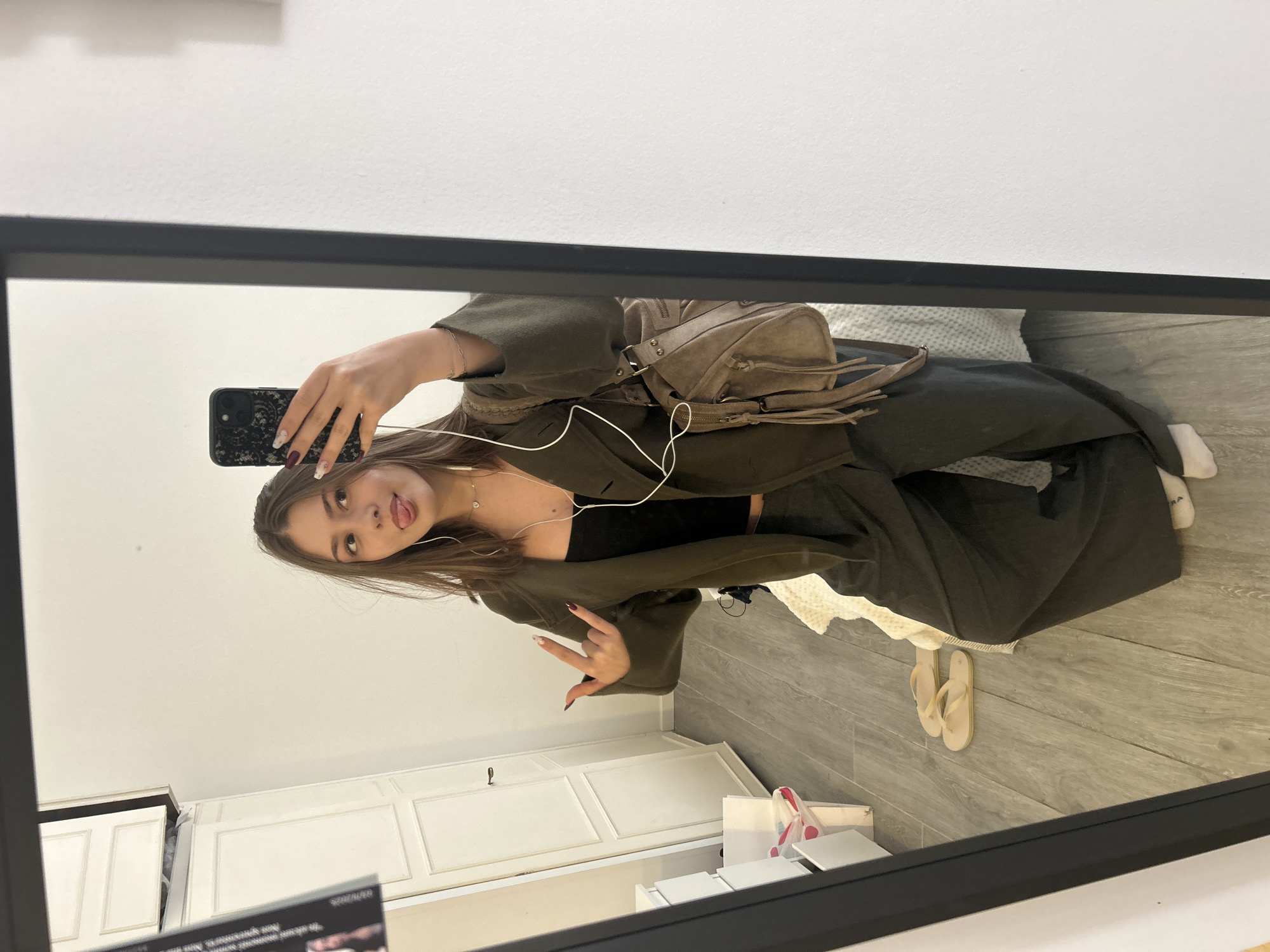 A young woman captures a mirror selfie in an indoor setting. The image, originally rotated 90 degrees, shows the woman reflected in a large rectangular mirror with a black frame. She has long brown hair and is wearing a dark olive green jacket or oversized shirt over a black top, with matching wide-leg pants. White wired earbuds are visible in her ears, connected to her black smartphone, which has a textured case and is held up to take the photo. She poses playfully, sticking her tongue out slightly and making a "rock on" or peace sign gesture with her left hand, revealing manicured nails with intricate designs.
Behind her, a light brown or beige suede backpack with fringe details and metal studs rests on what appears to be a bed or seating area, covered with a textured white blanket. The room features white walls and light gray/brown wooden flooring. To her left, a white cabinet or dresser with paneled doors and dark handles is visible. On the floor, a single white sock is seen on her right foot, and a pair of light-colored flip-flops are placed nearby, next to a white shopping bag with pink dots. The lighting is bright and even, suggesting daytime.
Faintly visible text in the bottom left reflection of the mirror includes what appears to be a date, "2023/10/30", and the Italian phrase "non sprecare" (don't waste), potentially from a label or sign. The overall scene suggests a casual moment, possibly in a home or hotel room in Milan, Italy.