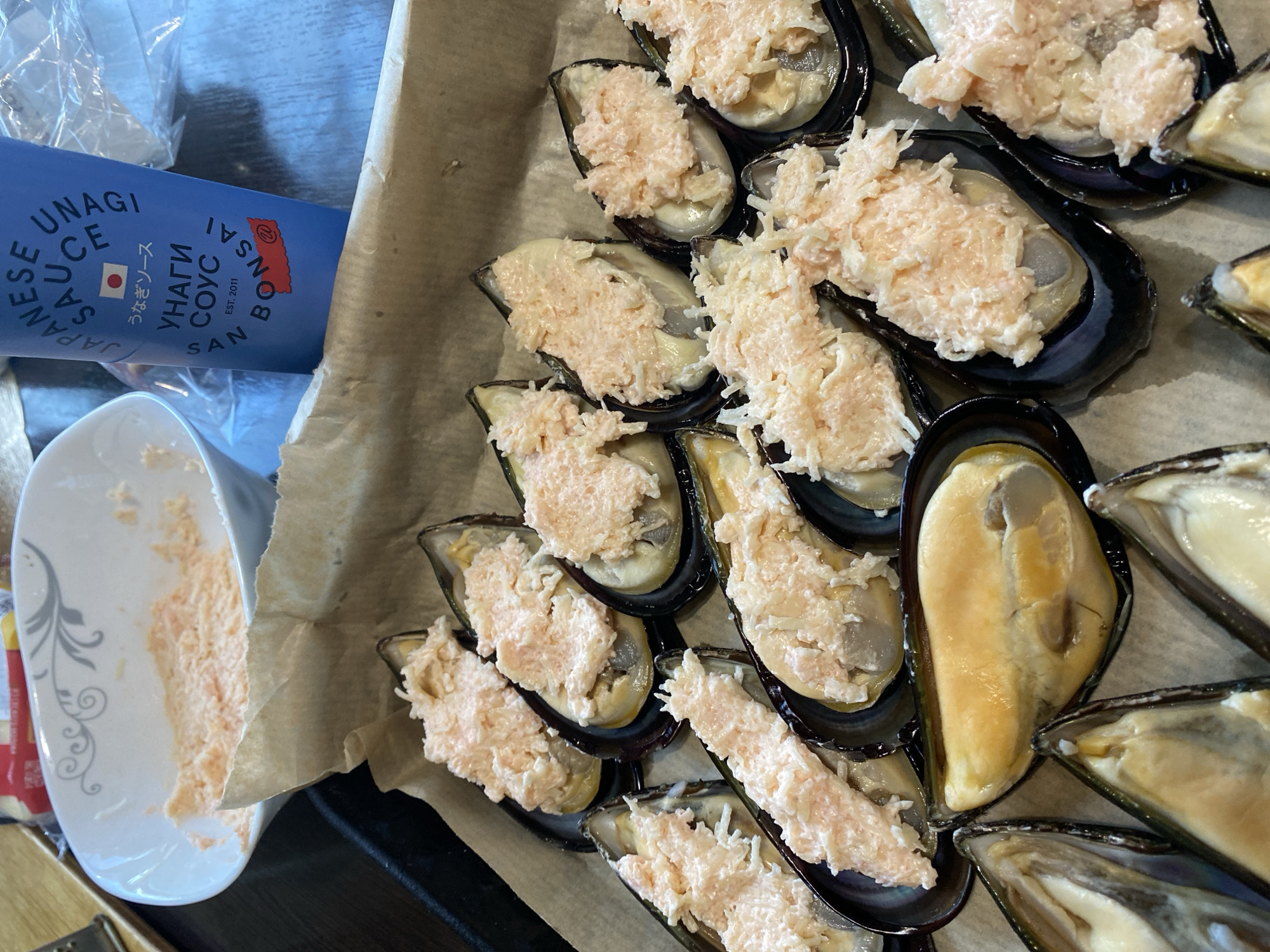 A close-up, high-angle shot shows a tray lined with parchment paper, filled with mussels that have been topped with a pinkish filling. The mussels are arranged in rows, with the filling spread across the visible half of each mussel. In the background, a white bowl contains more of the pinkish filling. To the right of the bowl is a blue bottle with Japanese text and an illustration of a red flag, likely containing sauce for the mussels. The overall scene suggests food preparation, with ingredients and tools laid out on a surface. The location is not determinable from the image.