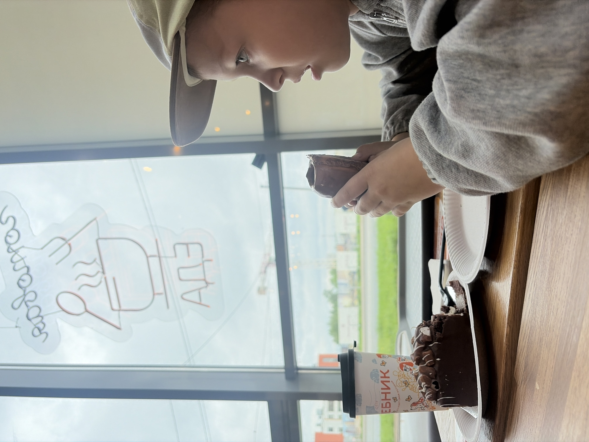 A young child wearing a baseball cap and gray sweatshirt is seated at a wooden table in an establishment. The child is holding a piece of chocolate pastry in their hands, possibly taking a bite or examining it. On the table, there is a slice of chocolate cake on a white plate, a plastic coffee cup with a pattern, and a fork. Through the window behind the child, a street and buildings are visible. A neon sign in Russian is affixed to the window. The location is Saint Petersburg, Russia.